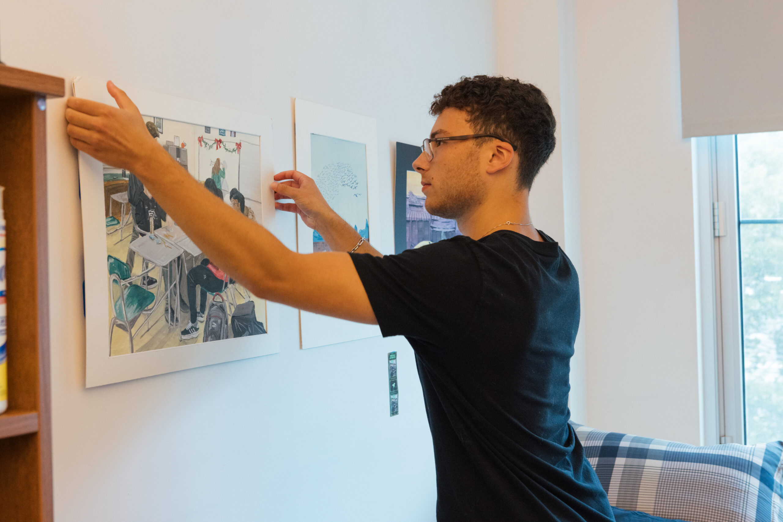 A young person with curly hair and glasses is hanging a framed artwork on a wall in a bright room. They are focused on positioning the artwork, which depicts a classroom scene. In the background, there is a blue and white checkered couch and a window letting in natural light.
