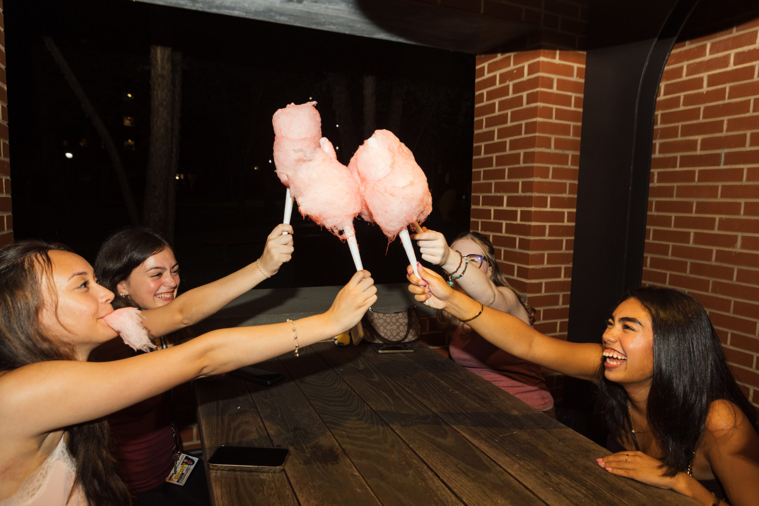 Four friends are smiling and enjoying cotton candy at a table. They are playfully raising their cotton candy sticks towards each other, with pink fluffy cotton candy on the ends. The setting is dimly lit, with brick walls and trees in the background, capturing a fun, social atmosphere.