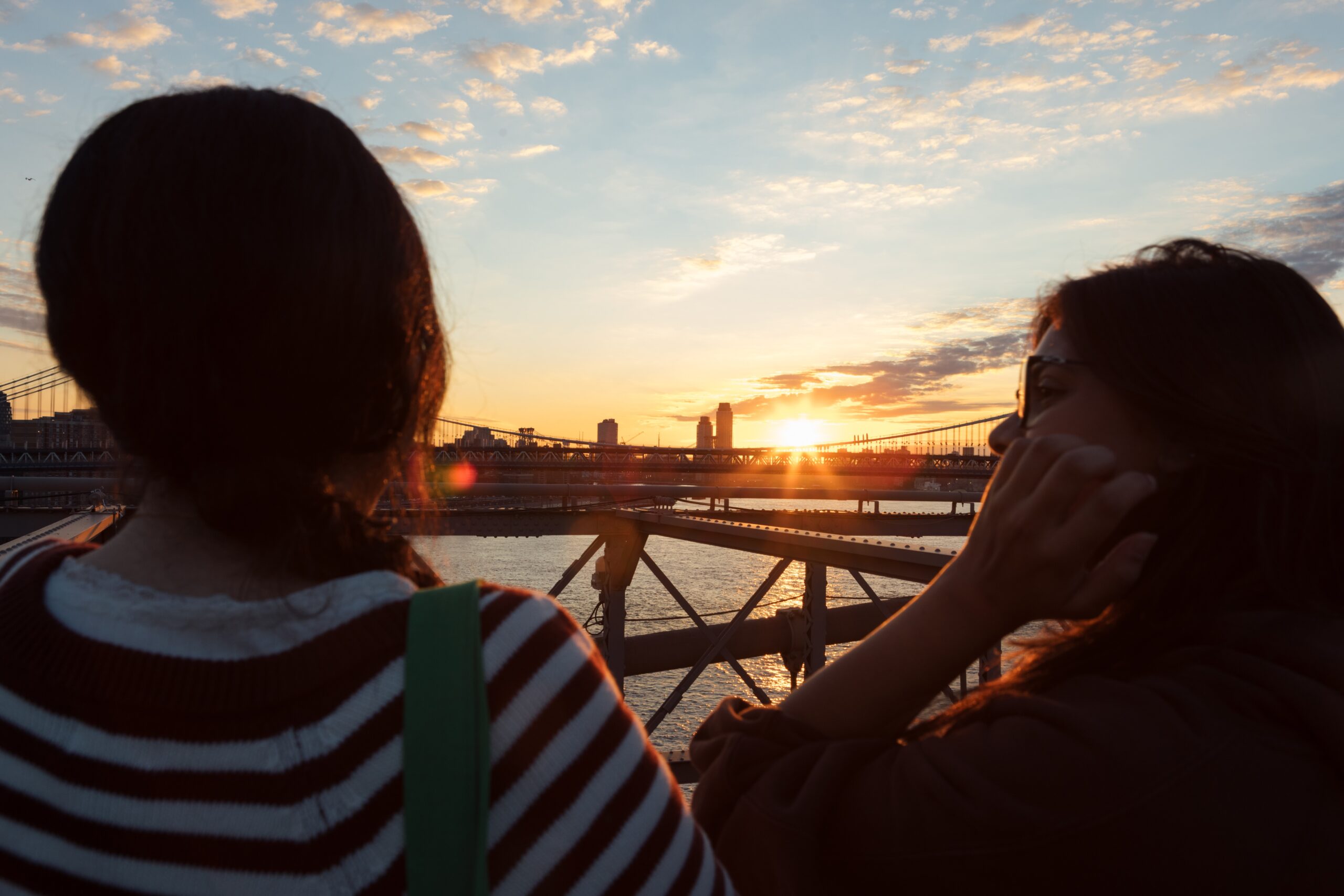Two individuals stand on a bridge facing a sunset over a river. The sky is painted with soft clouds in shades of orange and blue. One person, wearing a striped sweater, has their hair in a braid, while the other, wearing glasses, rests their hand on their face, deep in thought. The background features a city skyline and a horizon slightly blurred by the setting sun's light.
