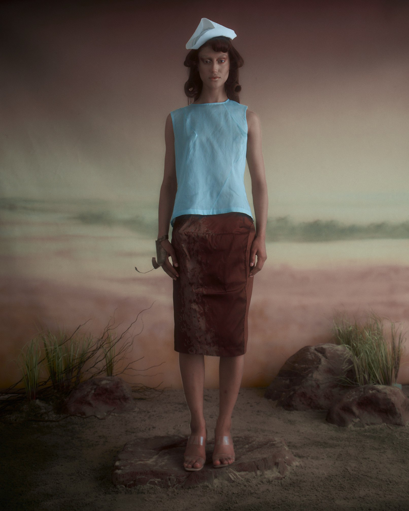 A model stands against a softly blurred background resembling a beach scene. She wears a light blue, sleeveless top and a brown skirt with subtle texture. Her hair is styled in loose curls, and she accessorizes with a white sailor hat. The ground is sandy with small rocks and tufts of grass, enhancing the natural setting. The atmosphere has a dreamy, ethereal quality.