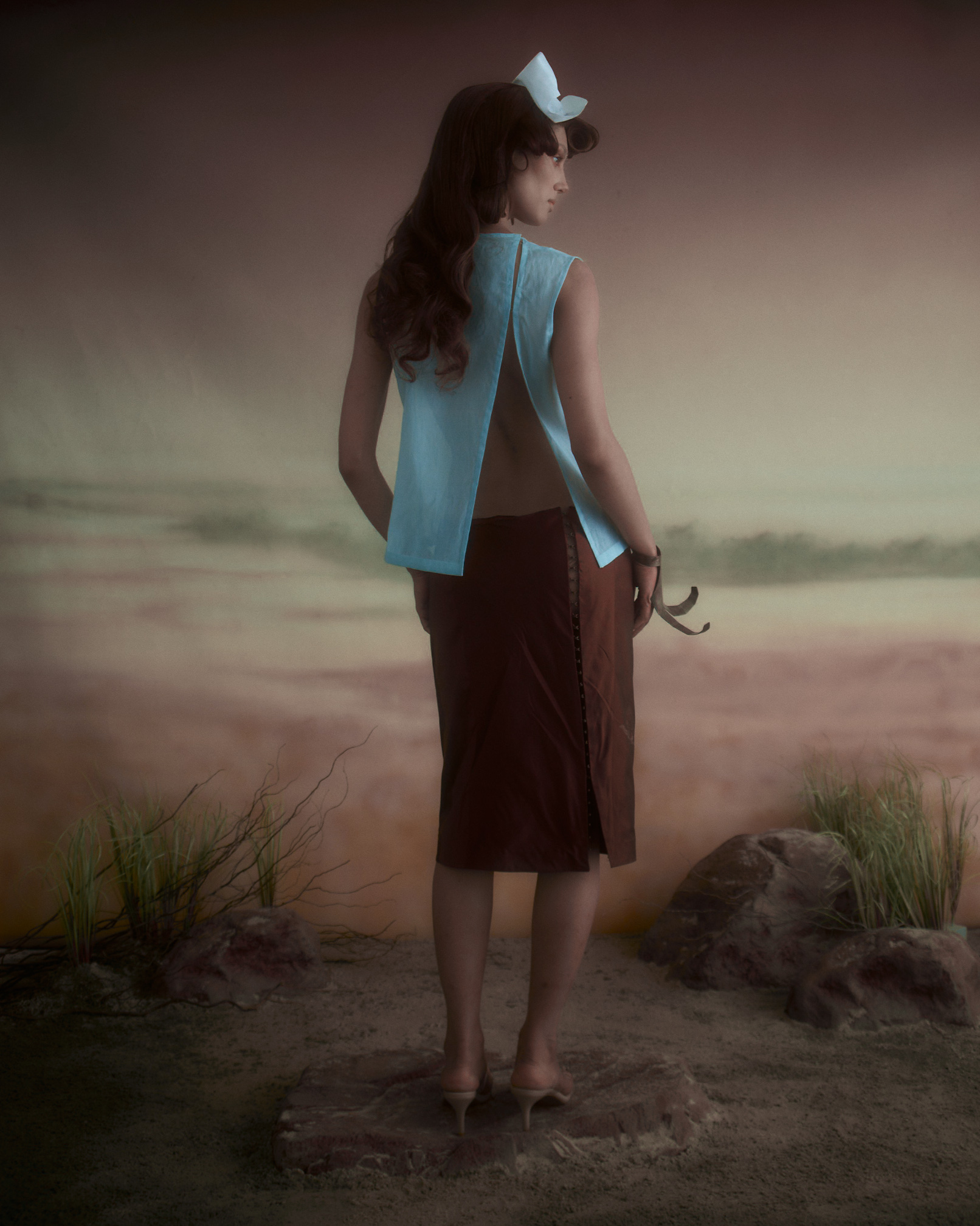 A model stands with their back facing the camera, wearing a light blue sleeveless top with an open back and a brown skirt. The scene has a soft, dreamlike background featuring sandy hues and abstract landscapes. The model has long, wavy hair and is wearing a white headpiece. The ground is textured, resembling a sandy surface with rocks and sparse vegetation.