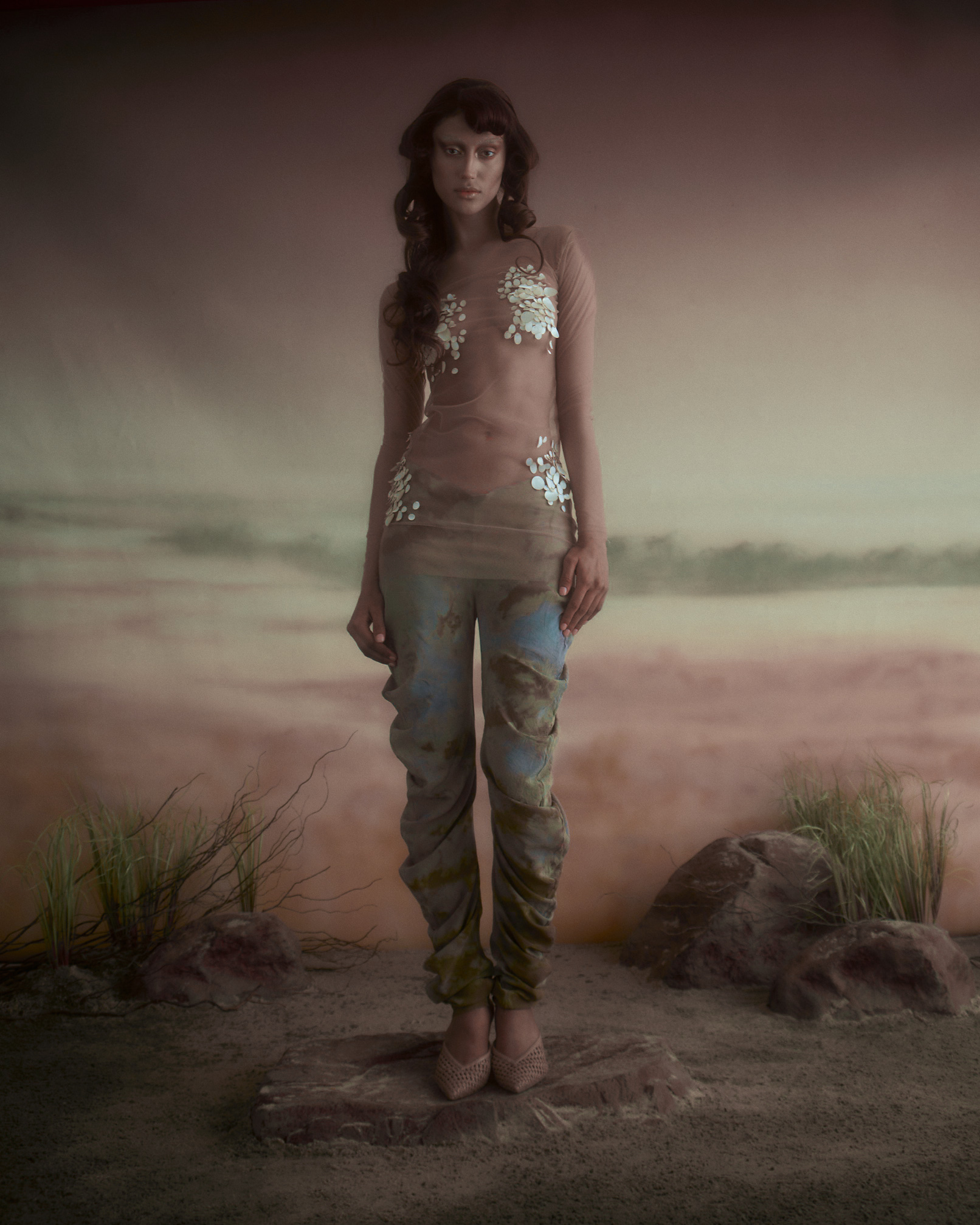A model stands on a rock in a surreal landscape, wearing a sheer top adorned with white floral designs and patterned blue-green pants. The background features soft, muted tones that resemble a sandy or earthy environment, with some grasses and rocks visible. The lighting is soft and ethereal, contributing to the dreamlike quality of the image.