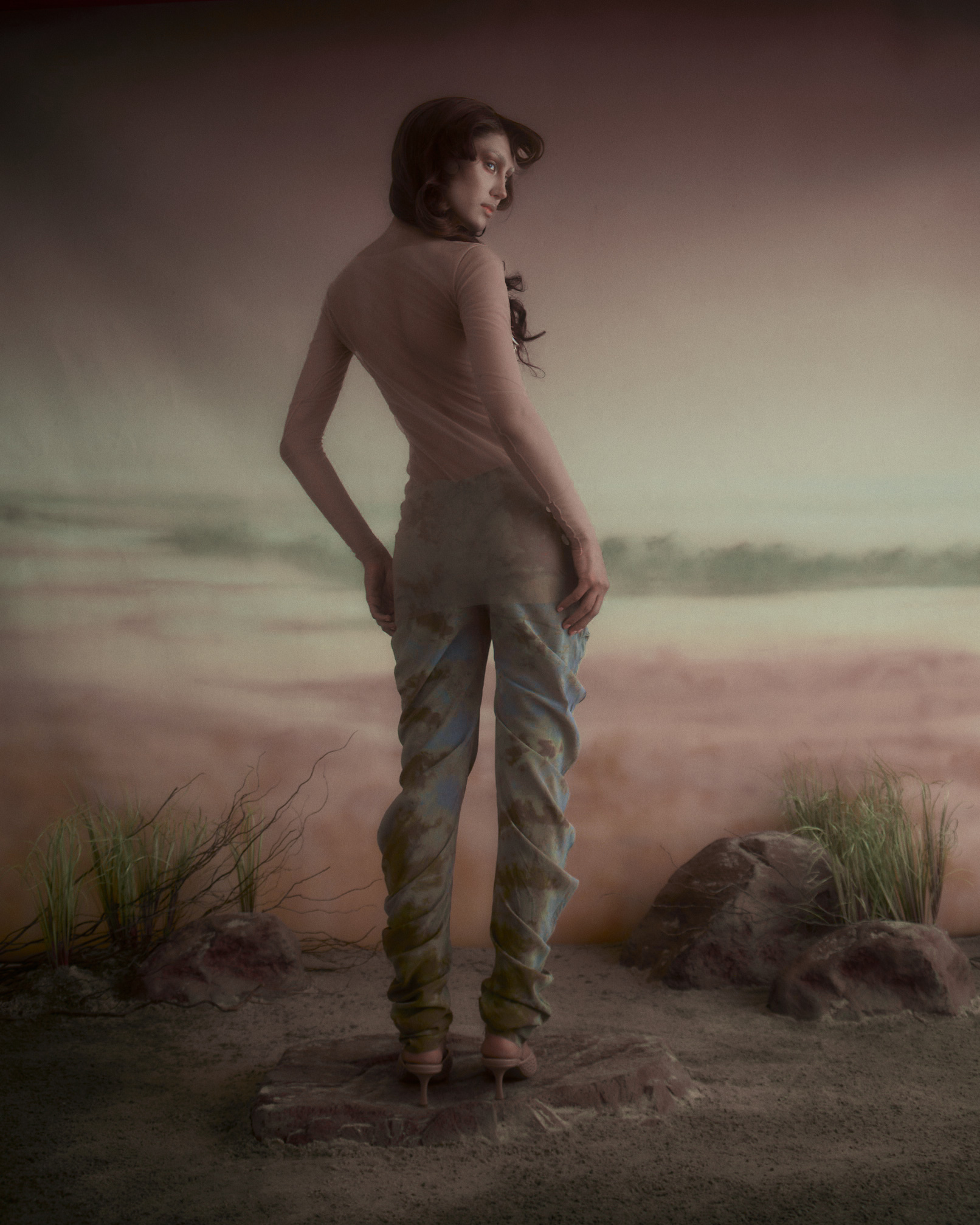 A figure stands with their back turned, wearing a fitted long-sleeve top and painted pants. The pants feature an abstract blue and green pattern, while the top is a soft pink. The setting includes sandy ground with scattered rocks and sparse grass, against a hazy, pastel-colored backdrop resembling a seascape. The figure's hair is styled in loose waves.