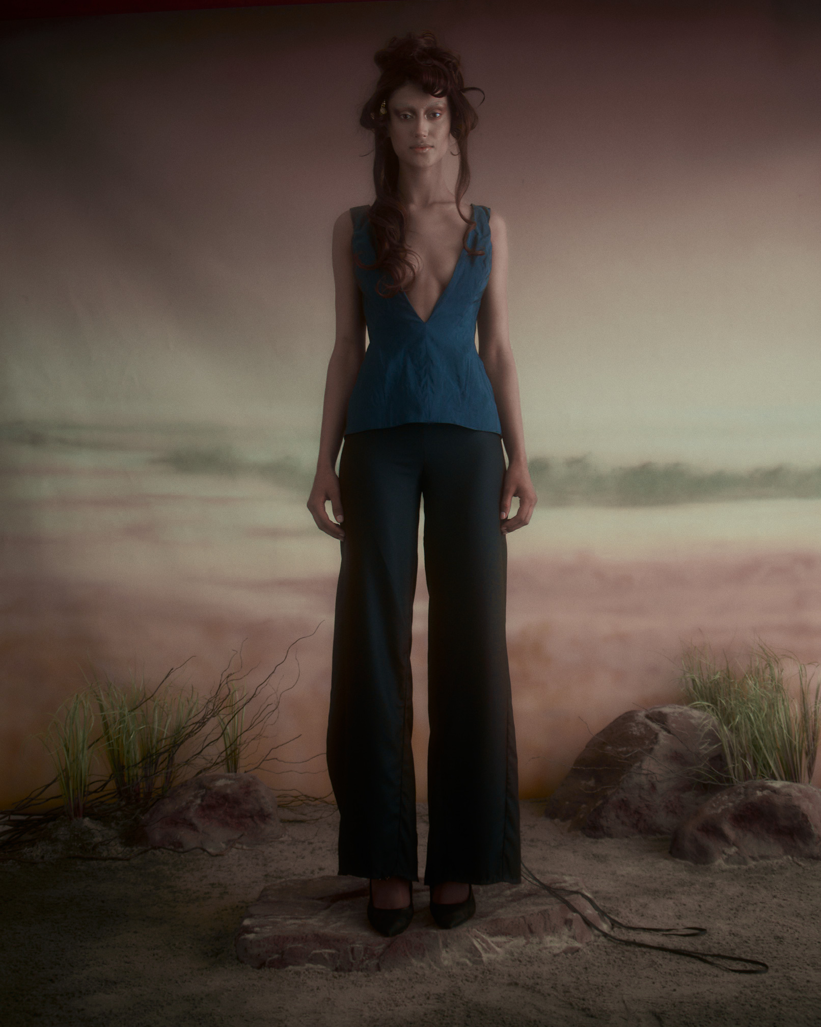A model stands in a surreal setting, wearing a deep blue top with a plunging neckline and black, wide-legged pants. The background features a soft, hazy landscape with muted colors and faint grass-like plants. The model's hair is styled in loose waves, and she has a minimal makeup look, conveying a dreamy aesthetic. The ground is textured with rocks and sandy elements, contributing to an artistic atmosphere.