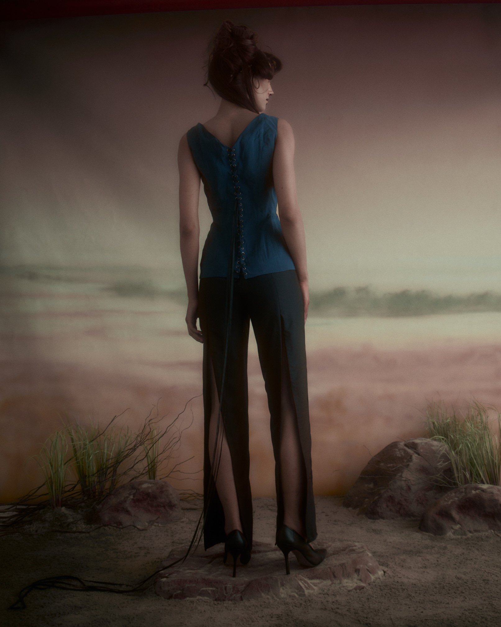 A model stands with her back to the camera, wearing a fitted blue top with a lace-up detail along the spine and high-waisted black pants featuring side slits. She is styled with an updo, and is situated against a softly colored background resembling a landscape, with abstract elements like grasses and rocks in the foreground. The overall mood is artistic and fashion-forward.