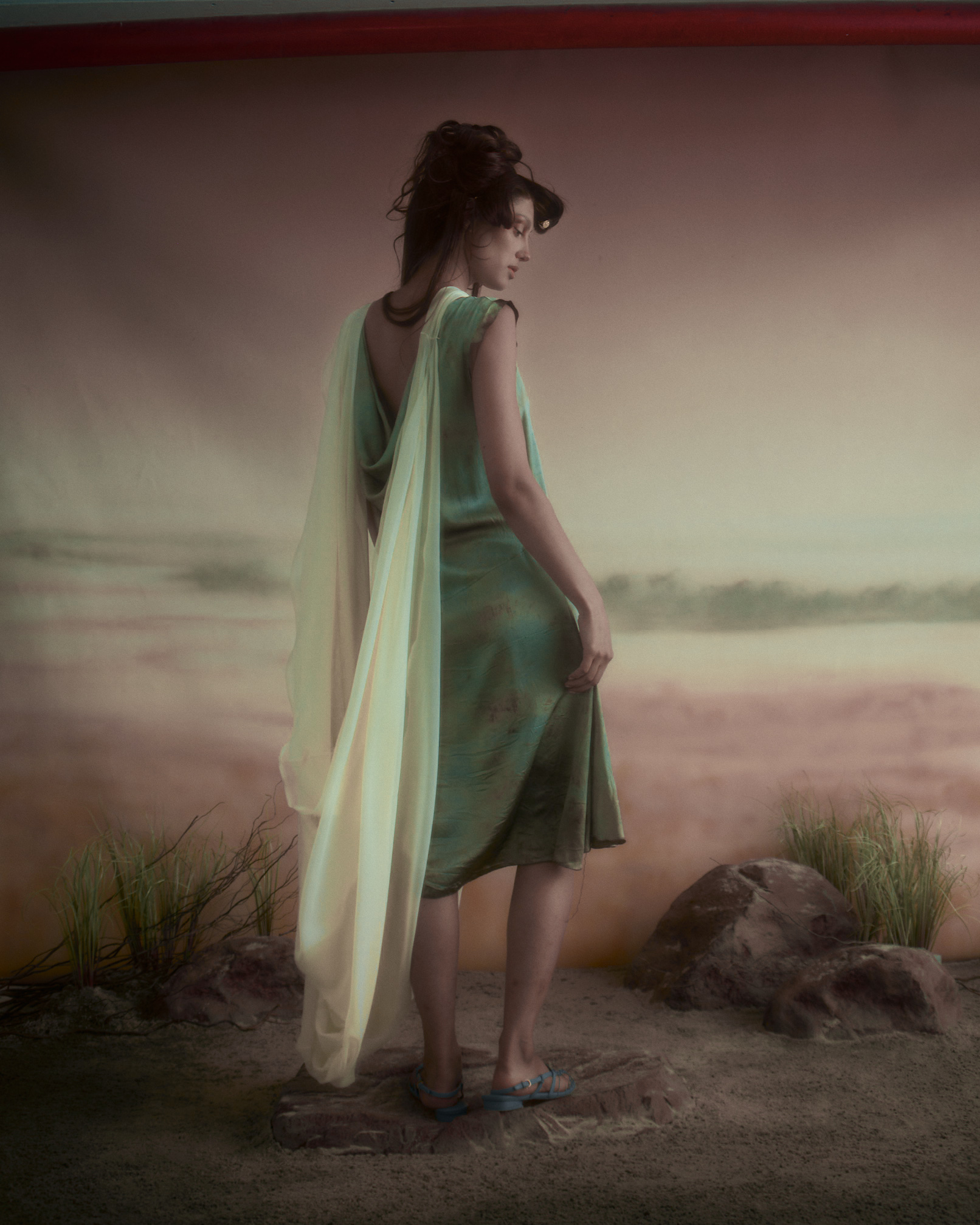 A woman stands with her back to the camera, wearing a green dress with flowing fabric draped over her shoulders. She has dark hair styled in loose waves and is positioned on a rocky base. The background features a beach scene with muted colors, suggesting a shoreline and grass. The overall mood appears serene and artistic.