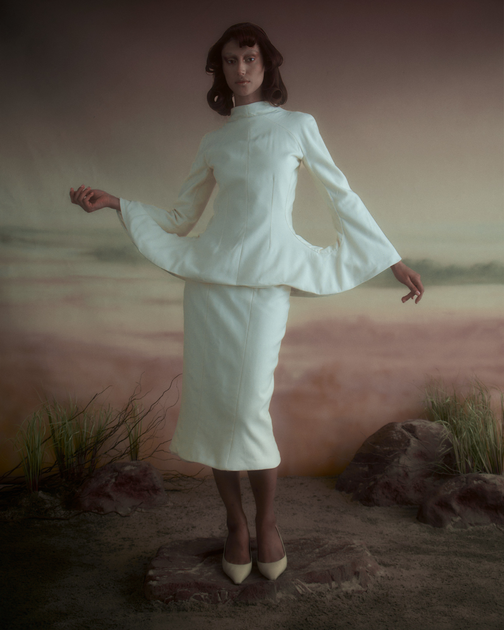 A model poses in a white, structured outfit with a high collar and bell sleeves, standing on a rocky surface. The background features soft, muted tones, resembling a surreal landscape. The model's hair is styled in soft waves, and she wears beige heels, creating an elegant and artistic impression.