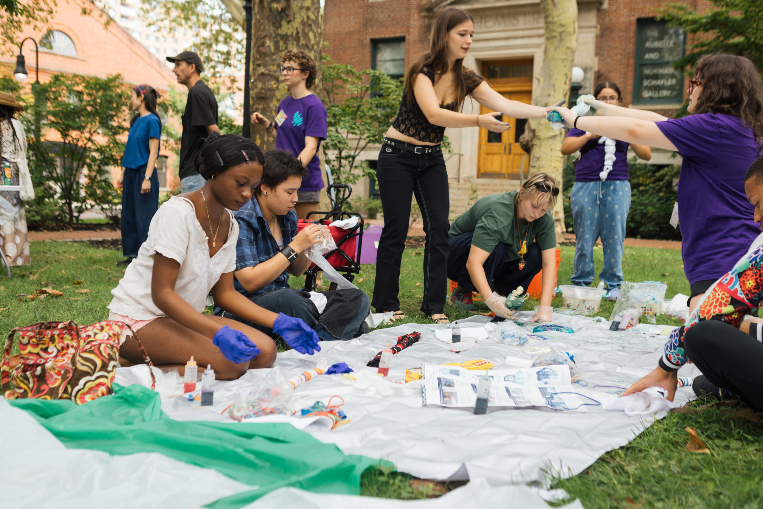 A group of diverse individuals are engaged in an outdoor creative activity. In the foreground, two people are focused on tying or dyeing fabric, surrounded by various materials like dye, rubber bands, and bottles. Others can be seen in the background, some standing and conversing, while others work on their projects on a large white tarp laid on the grass. The atmosphere appears collaborative and artistic, with trees and a building visible nearby.