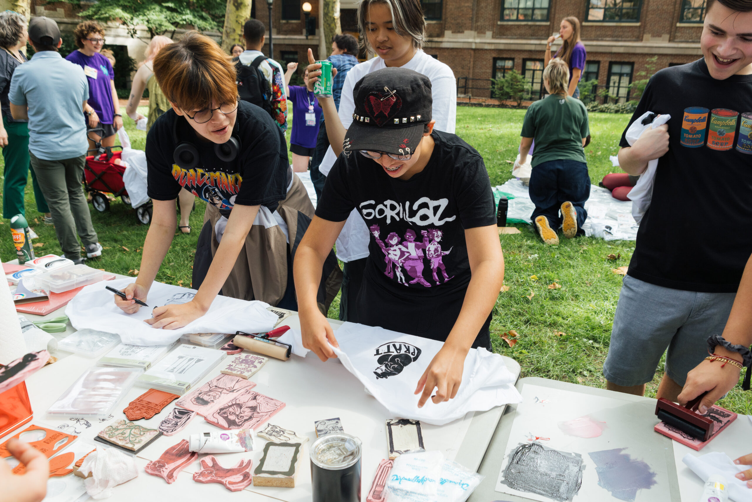 A group of young people is engaged in creating designs on T-shirts at an outdoor event. One person wears a black Gorillaz shirt while another focuses on a white T-shirt laid out on the table. Various art supplies, including stamps and paints, are scattered across the table. Others in the background are participating, with some seated and others standing, showcasing a lively atmosphere.