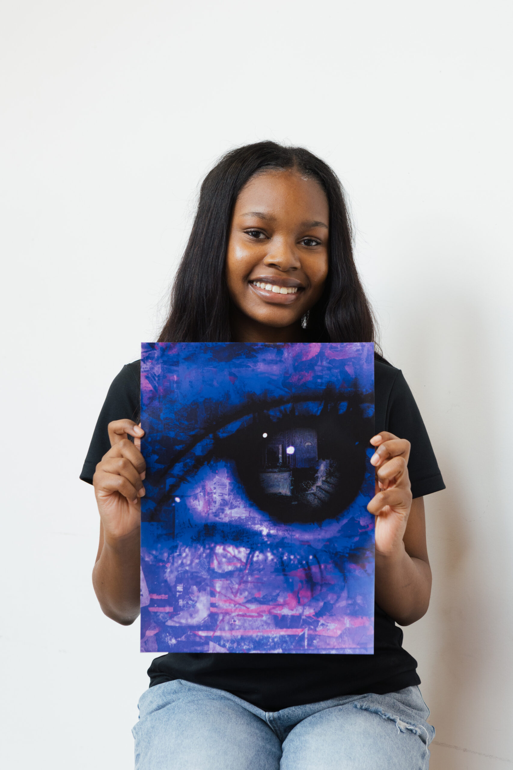 A young woman with long, dark hair is seated against a white background, smiling and holding a colorful poster featuring a large, artistic eye design in shades of blue and purple. She is wearing a black t-shirt and light blue jeans.