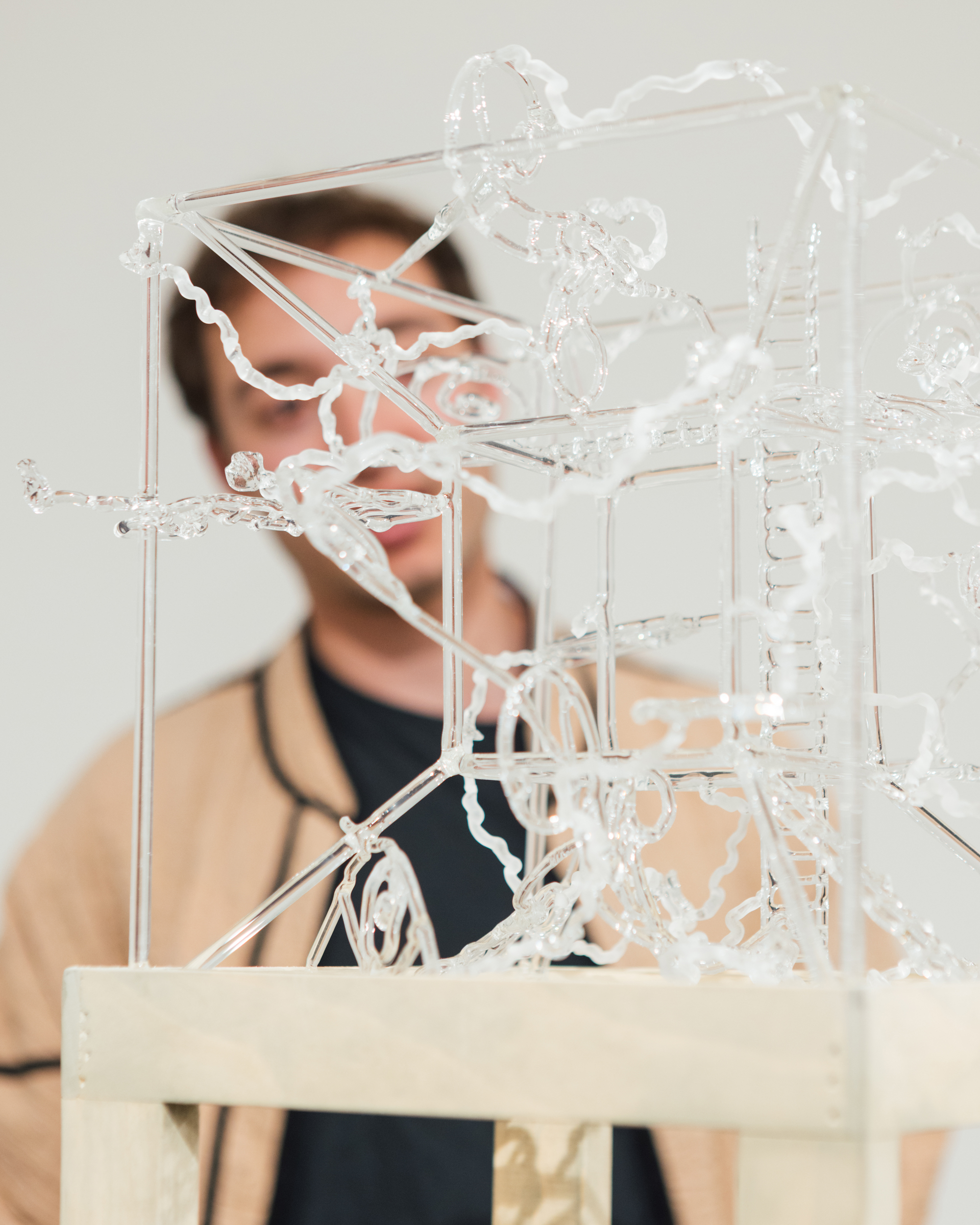A translucent glass sculpture with intricate, winding shapes is displayed on a wooden base. In the background, a person is partially visible, slightly out of focus, wearing a black shirt and a tan jacket, observing the artwork. The overall setting appears to be an art gallery or exhibition space with a plain backdrop.