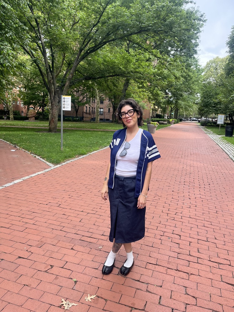 A person stands on a brick pathway surrounded by green trees. They are wearing a navy and white short-sleeved top over a white shirt and a knee-length denim skirt. Their feet are adorned with white socks and black shoes. The background features a park-like setting, with signs and a pathway leading into the distance.