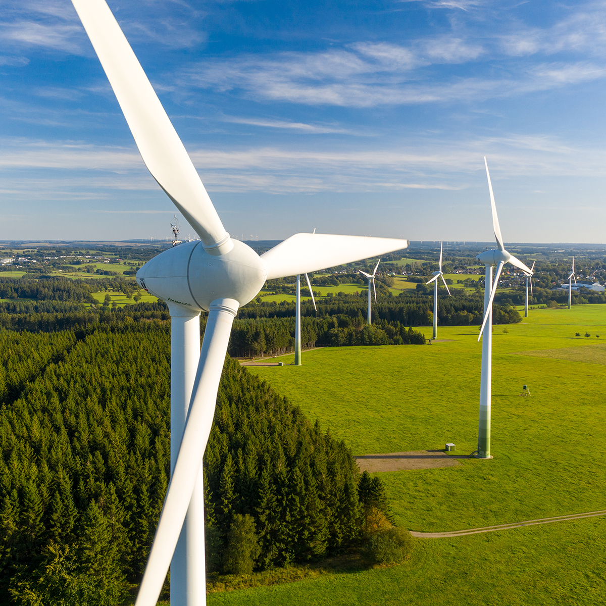 Aerial view of several white wind turbines situated in a green landscape. The foreground shows a close-up of one turbine's blades and hub, while in the background, more turbines are lined across a horizon featuring open fields and scattered trees. The sky is clear with a few clouds, indicating sunny weather.