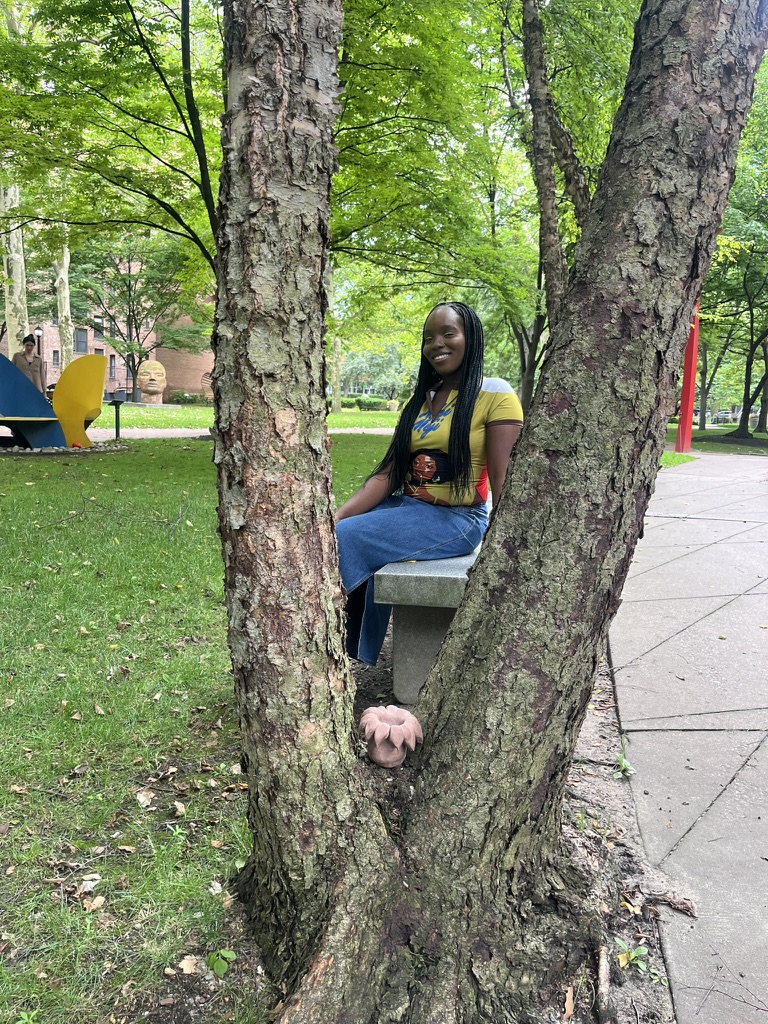 A person with long black hair, wearing a yellow shirt and blue jeans, smiles while sitting on a stone bench between two trees. The trees have textured bark and greenery surrounds them, creating a natural, park-like setting. In the foreground, there is a soft, round object resembling a mushroom at the base of one tree. In the background, colorful playground equipment can be seen.