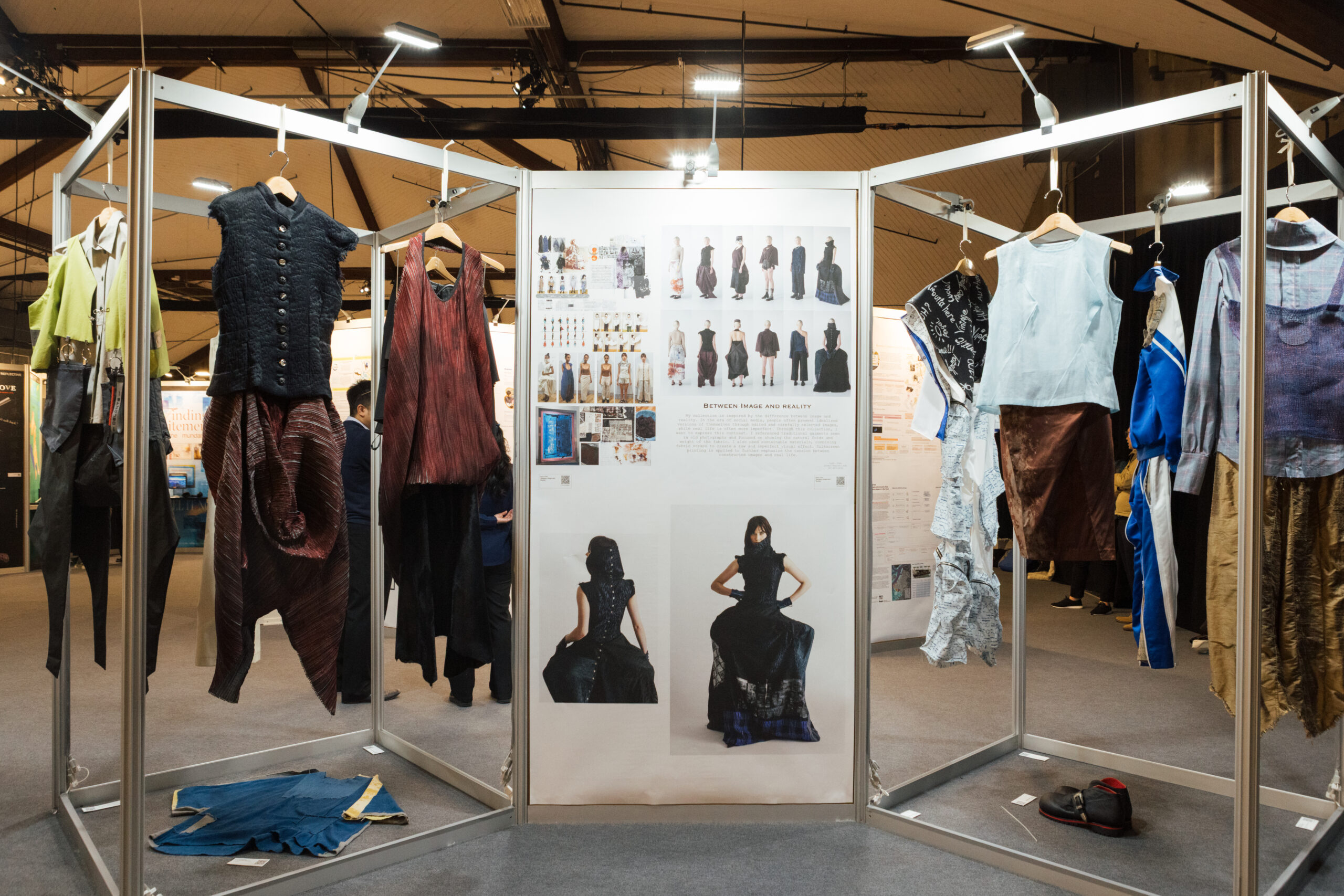 A fashion exhibition display featuring various garments hanging on metal frames. Prominent pieces include a black vest with buttons, a red flowing top, and several other unique clothing items, all displayed against a backdrop of design sketches and images of models wearing the styles. The arrangement suggests a theme of exploration between image and reality in fashion.