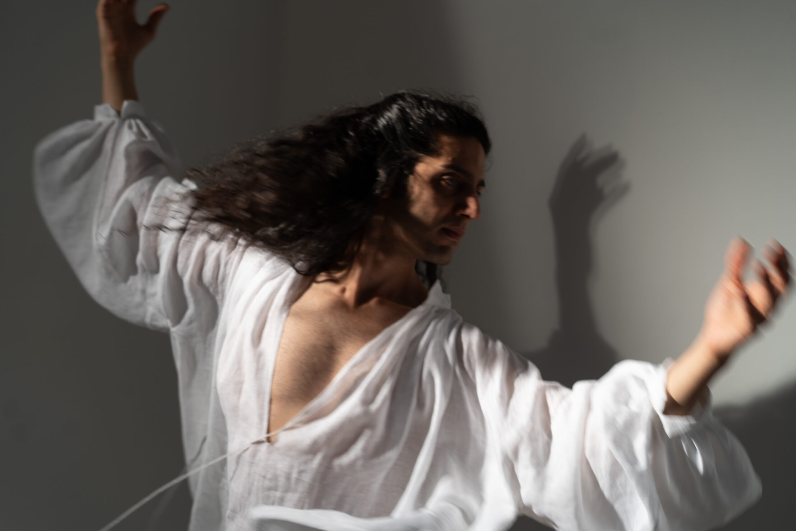 A dancer in a flowing white shirt is captured mid-movement, with long, dark hair cascading around. The dancer's posture and gestures convey emotion, and shadows on the wall add depth to the scene, emphasizing the dynamic nature of the performance. The background is a soft gray, enhancing the focus on the figure.
