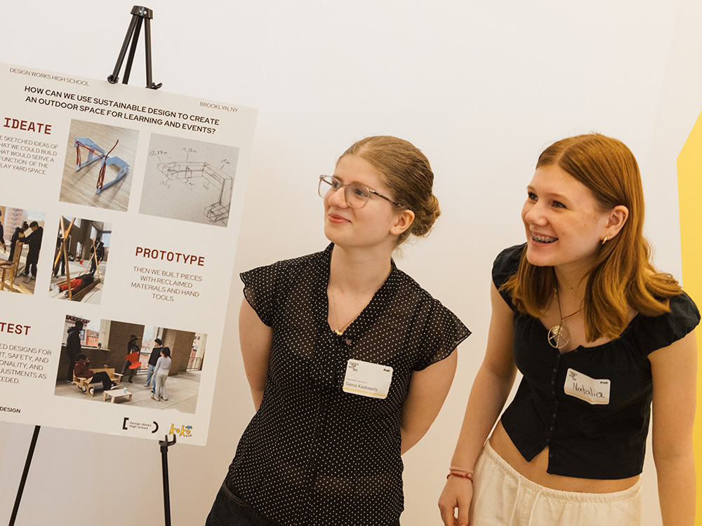 Two young students stand next to a poster discussing sustainable design for creating an outdoor space for learning and events.
