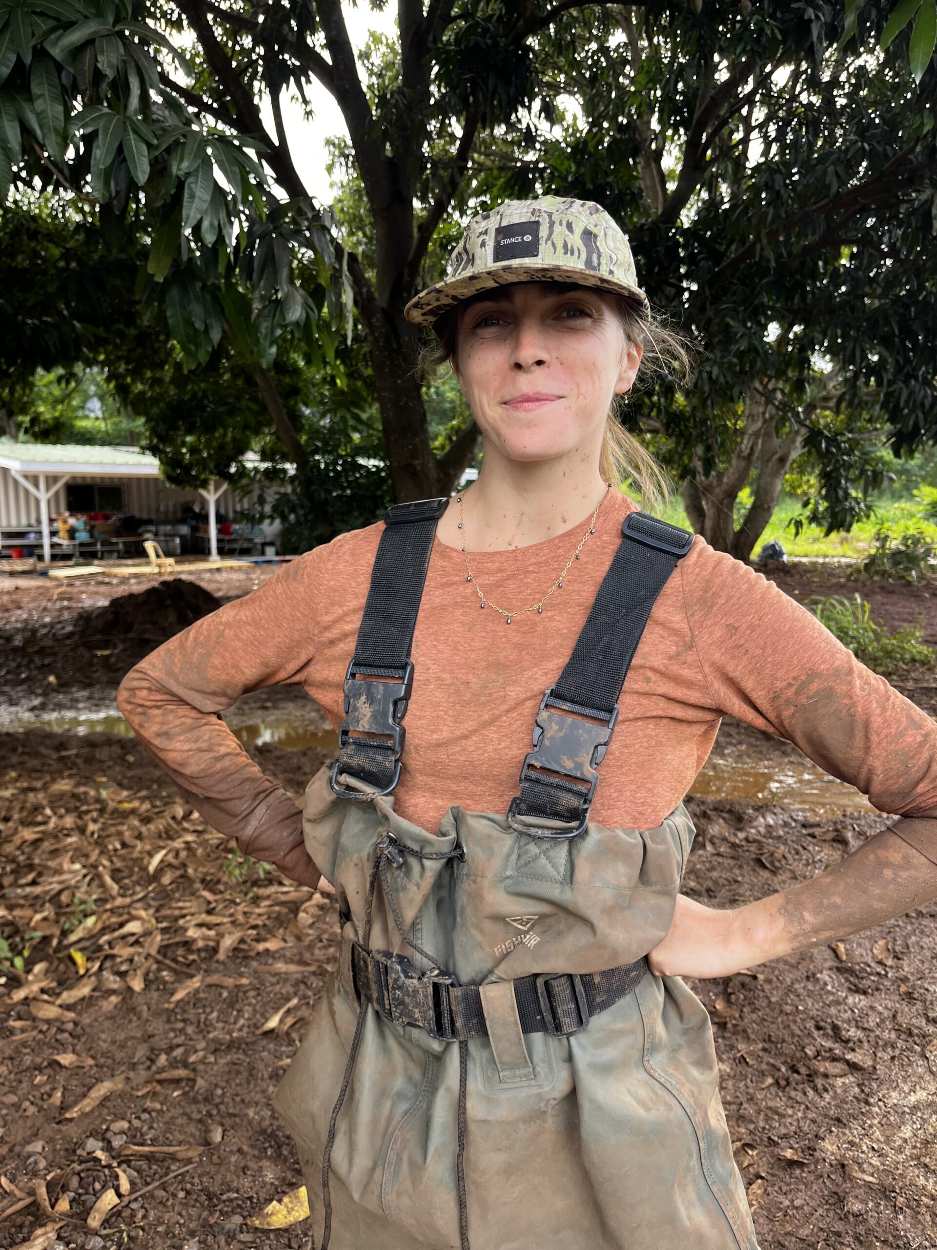 A woman stands outdoors wearing a camouflage cap and a long-sleeve rust-colored shirt. She has muddy arms and is dressed in green waders with straps. The background features lush greenery and a partially visible structure. The scene conveys an active, outdoor environment, likely related to a farming or gardening activity.