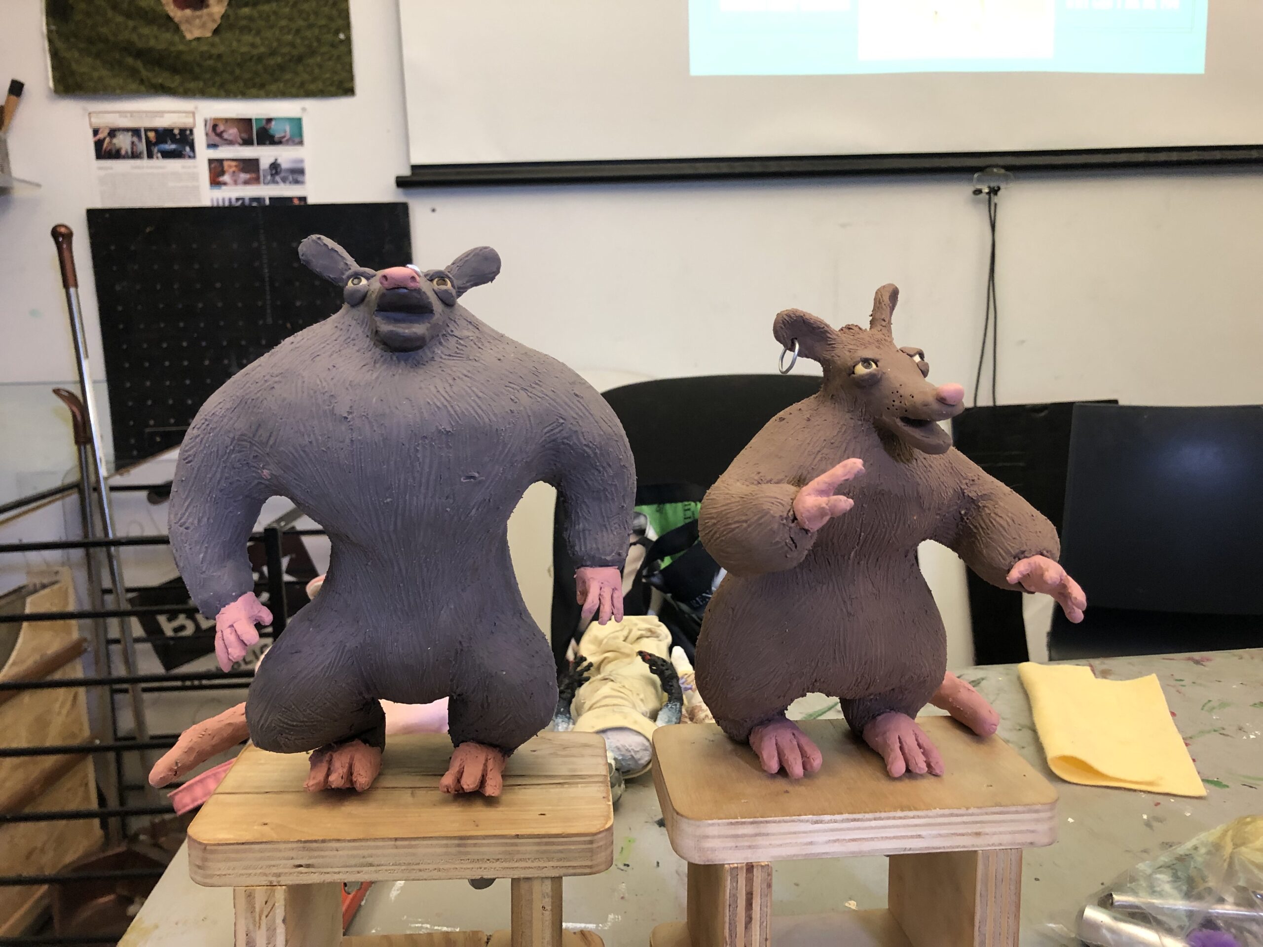 Two clay figures resembling anthropomorphic rats stand on wooden bases. The left figure has a muscular build and is painted gray, while the right figure appears more rounded and is painted brown. Both figures have detailed facial features, including ears and pink accents on their hands and feet. In the background, there are various art supplies and a projector screen.