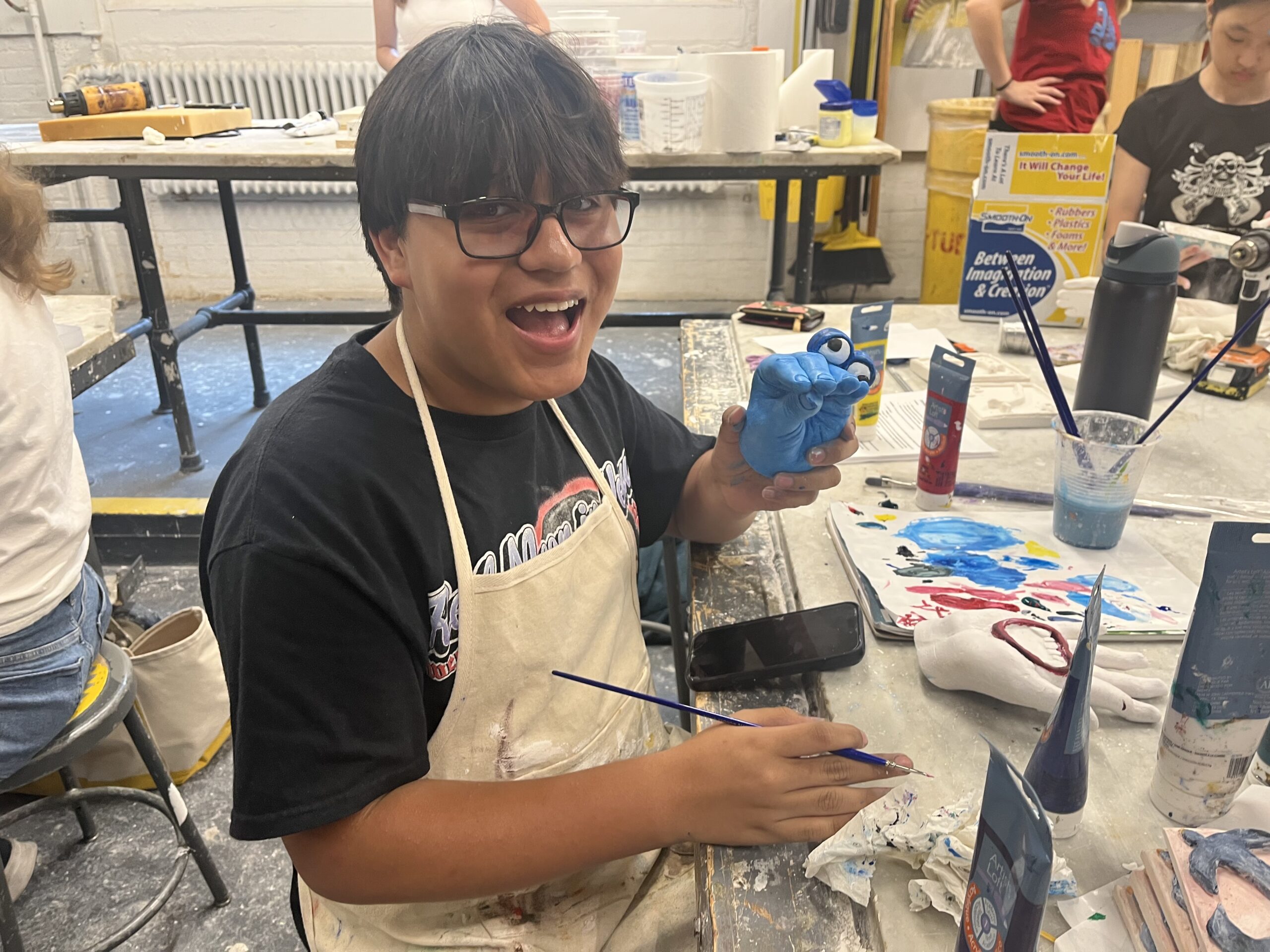 A young person with dark hair and glasses is sitting at a craft table, smiling and holding a blue, clay figure with eyes. They are wearing an apron over a black t-shirt and have paintbrushes and paint tubes in front of them. The background shows other people working and a variety of art supplies scattered on the table.