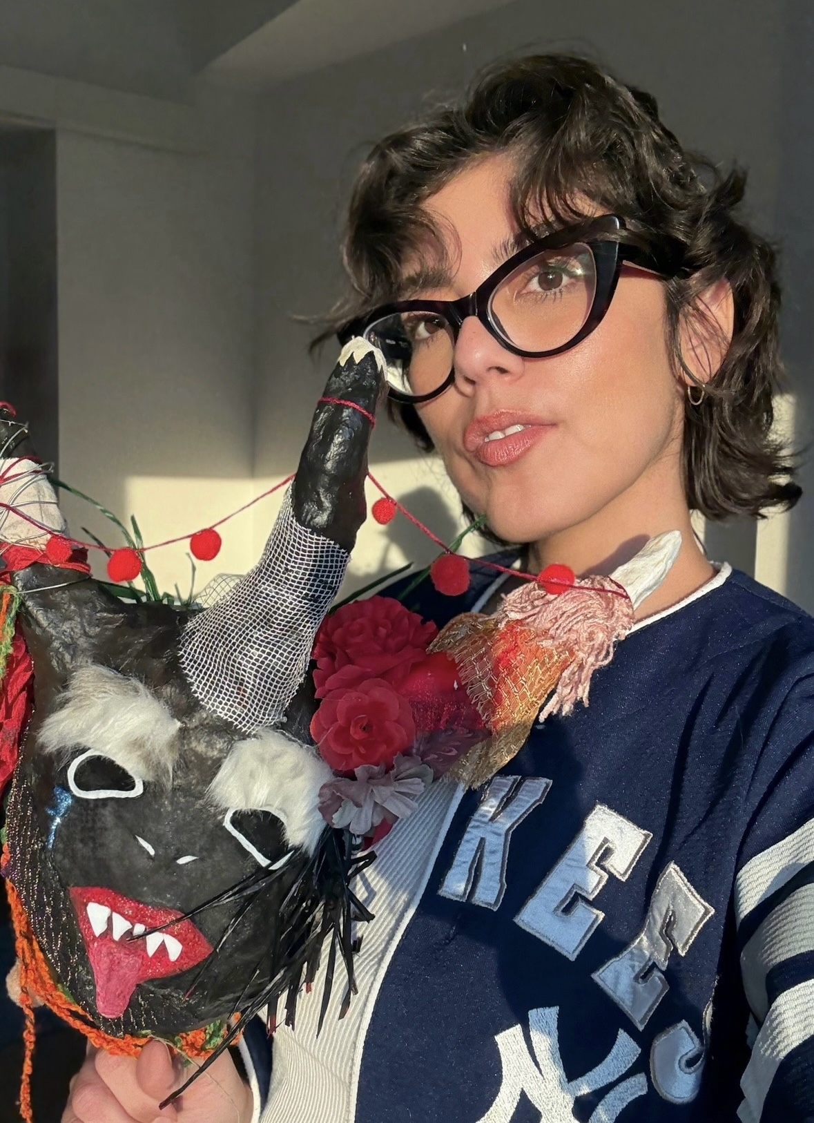 A person with short, curly hair, wearing oversized glasses, poses for a selfie while holding a colorful, artistic mask. The mask features exaggerated facial features, including sharp teeth and detailed decorations like flowers and ribbons. The background has soft lighting, suggesting a cozy indoor setting. The person has a slight smile, engaging with the camera.