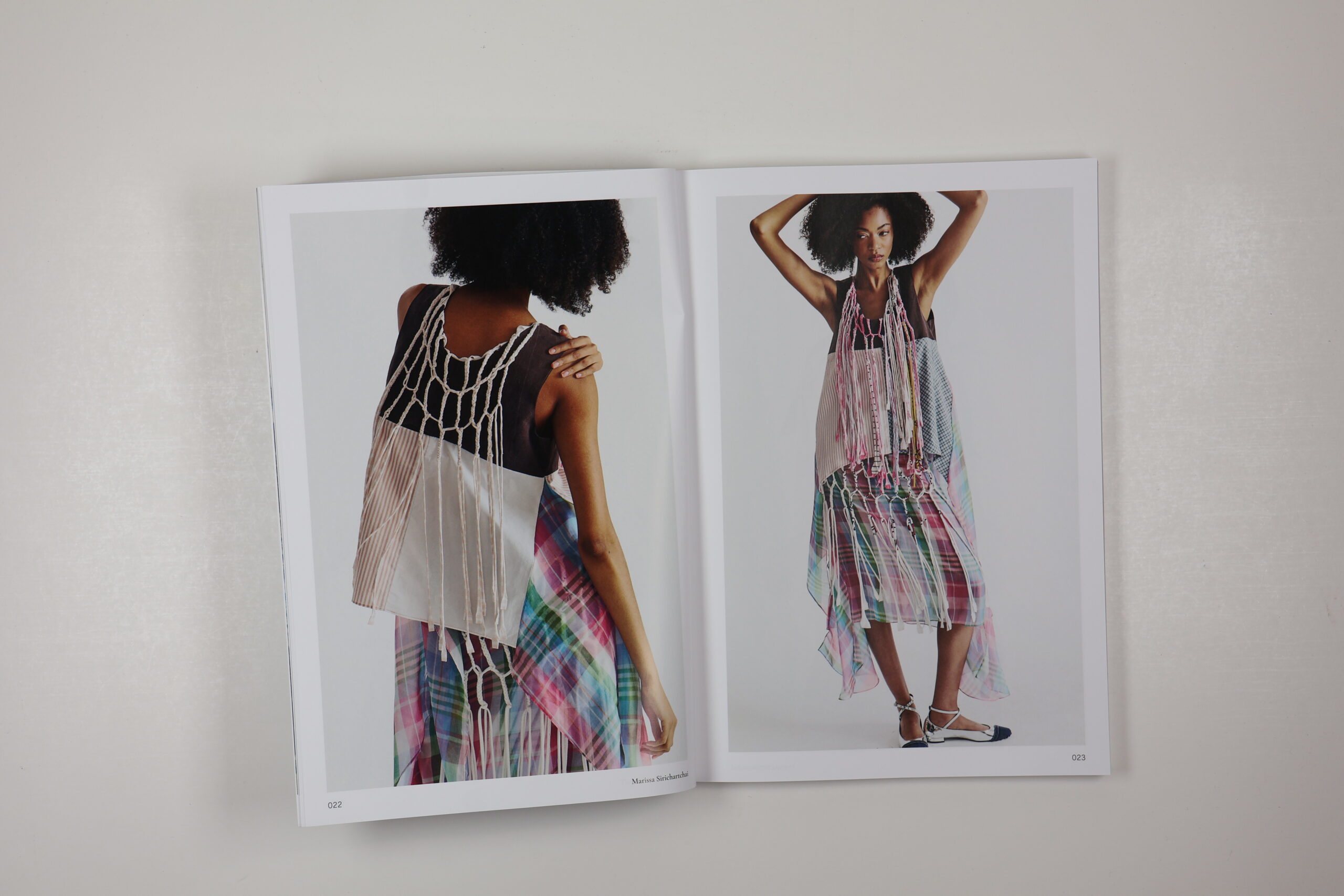 A fashion magazine spread features a model wearing a unique, layered outfit that combines various patterns and textures. The left page shows the back view, highlighting a macramé design and a mix of solid and sheer fabrics. The right page displays the front view, where the model poses with arms raised, showcasing the asymmetrical cut and colorful plaid patterns of the garment. The pages are numbered 022 and 023.