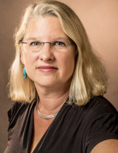 A woman with shoulder-length blonde hair is looking directly at the camera. She wears glasses and has turquoise earrings. Her outfit is dark with a V-neck style, and she has on a silver necklace. The background is softly blurred in warm tones.