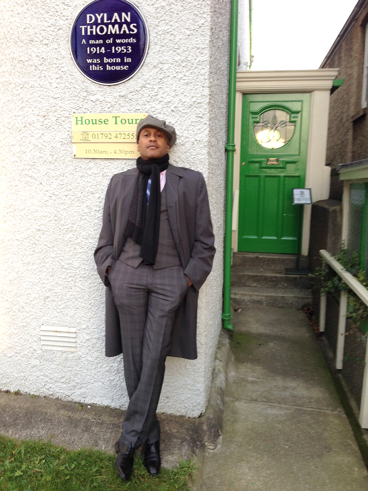A person in a stylish gray outfit and hat stands beside a white wall with a blue plaque that reads "DYLAN THOMAS A man of words 1914-1953 was born in this house." There is a green door to the right and a sign below indicating house tour details. The background features a small garden area.