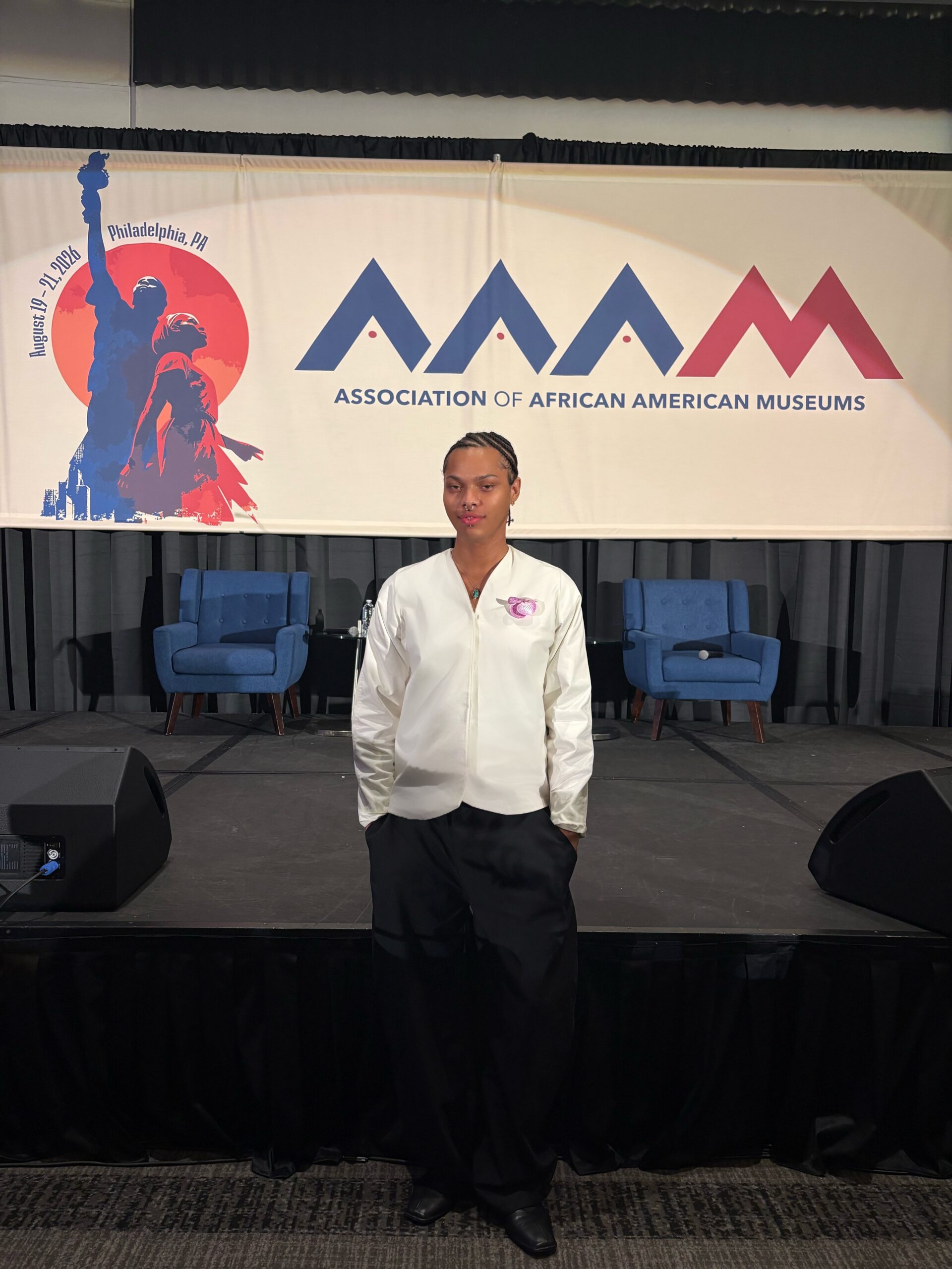 A person stands in front of a large banner for the Association of African American Museums, which features a graphic of a statue and text about the event taking place in Philadelphia, PA, from August 19-21, 2023. The individual wears a white shirt and black pants, with their hands in their pockets. Behind them is a stage with two blue chairs.