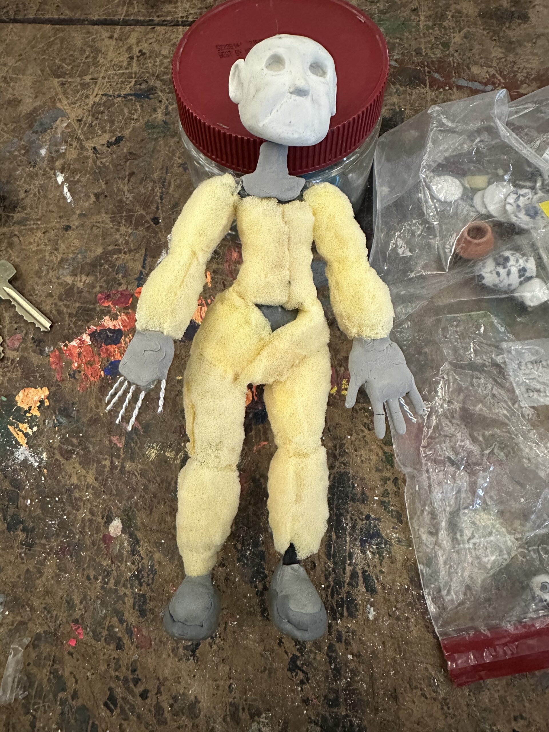 A handmade doll with gray hair and a painted face is being held by a person. The doll wears a white dress with a red trim and a green felt cape secured with a green button. The background features a black cutting mat and some scissors, indicating a crafting environment.