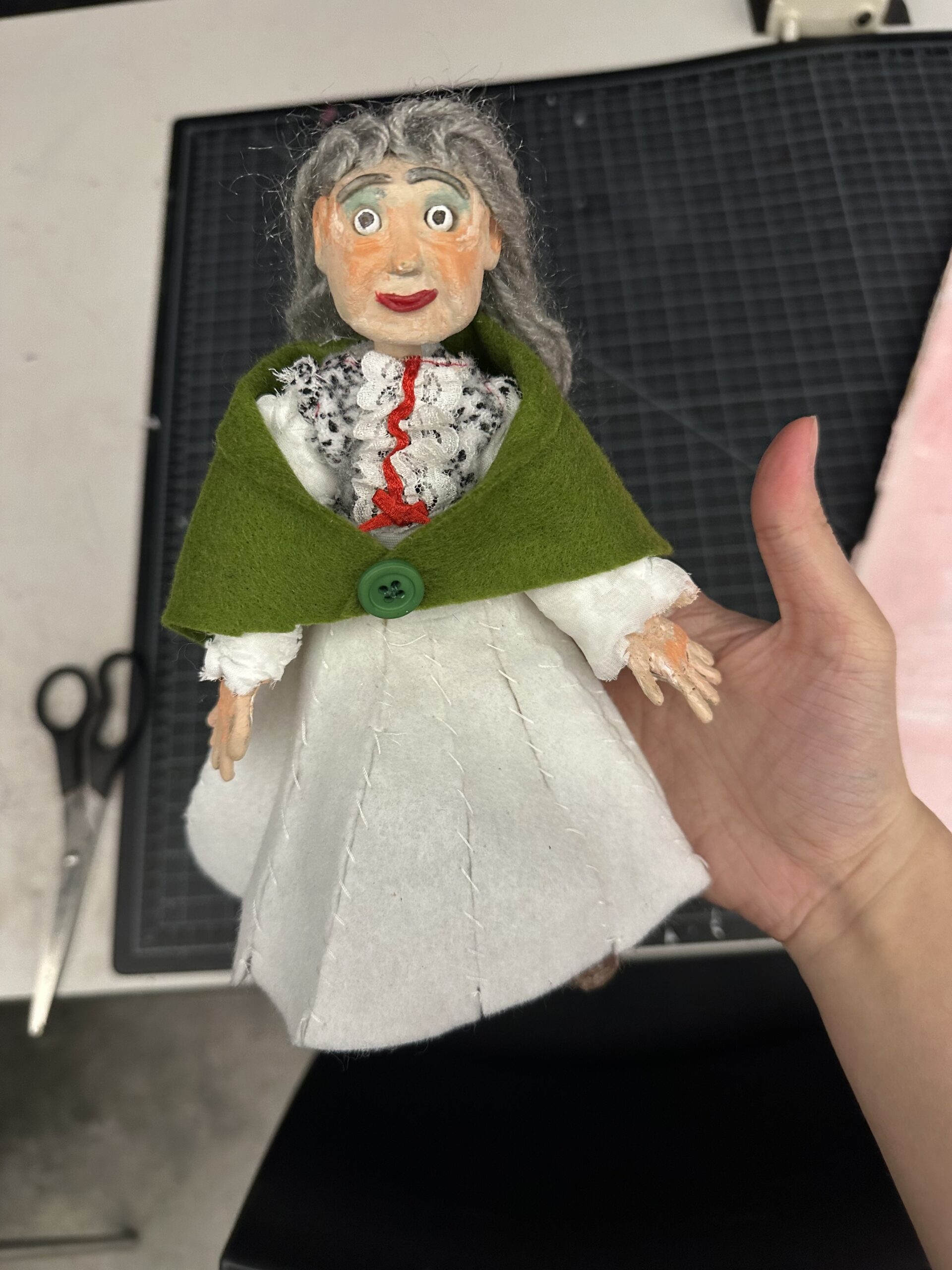A hand holds a handmade doll with a painted face, gray hair, and a green felt cape. The doll wears a white dress with red and black decorative elements. In the background, there are scissors and a cutting mat, indicating a crafting environment.
