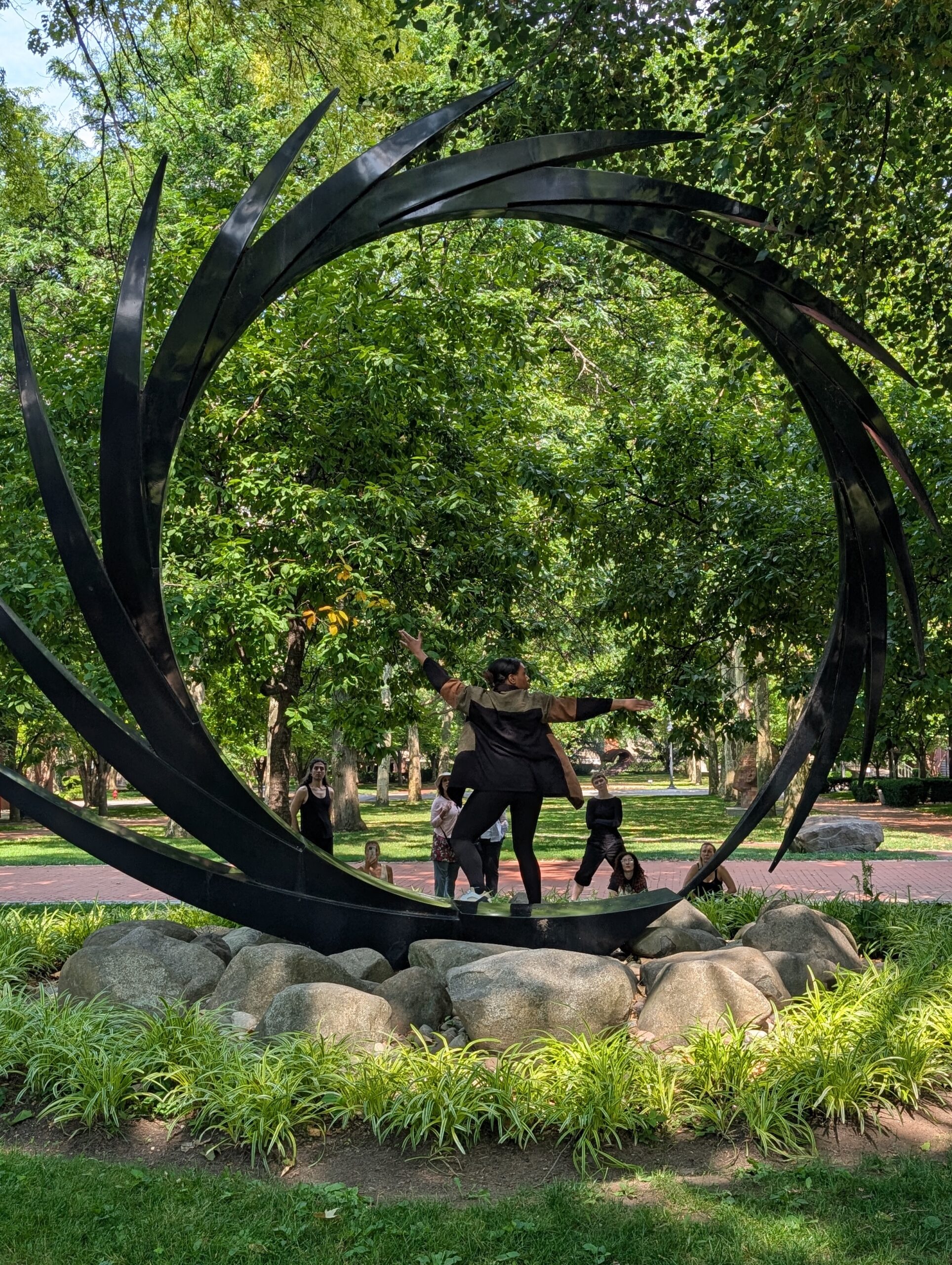 A person in a black outfit poses dramatically on a stone pedestal, centered in a circular black sculpture with protruding spikes. She appears to be performing or striking a pose. In the background, several people watch and take photos, surrounded by lush green trees and grass in a park setting.