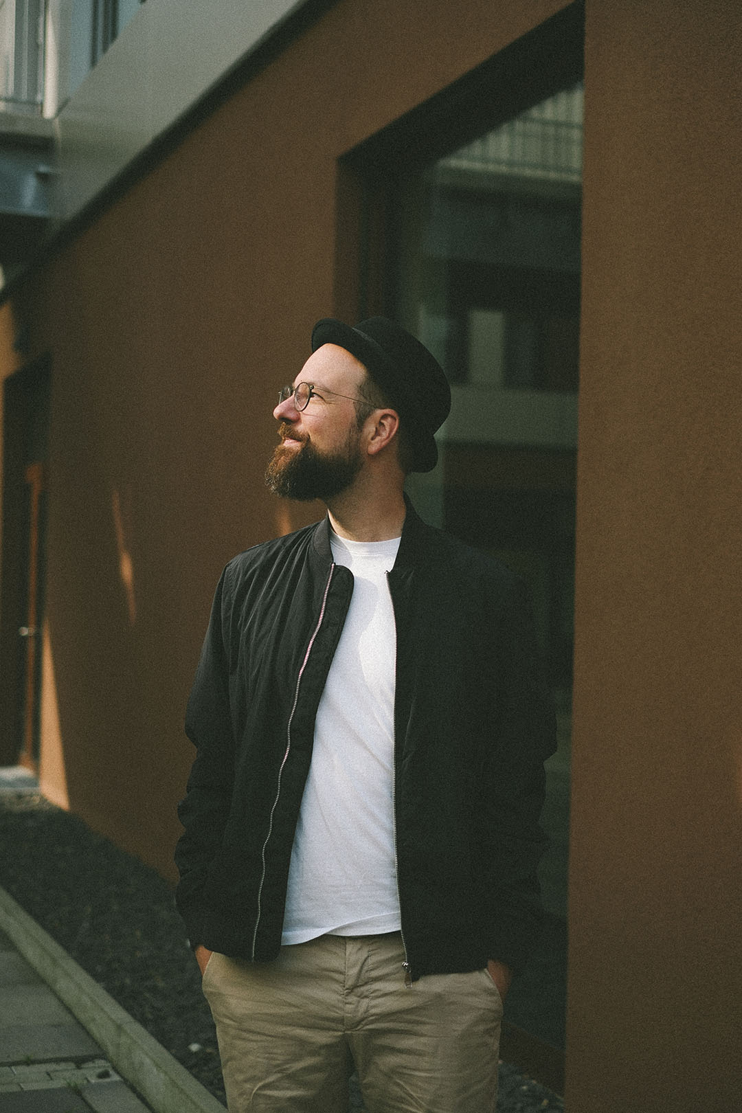 A man with a beard and glasses stands outside against an orange wall, wearing a black bomber jacket and a white t-shirt. He has a black hat and is looking up, appearing contemplative. Paved stones and dark pebbles are visible at his feet.