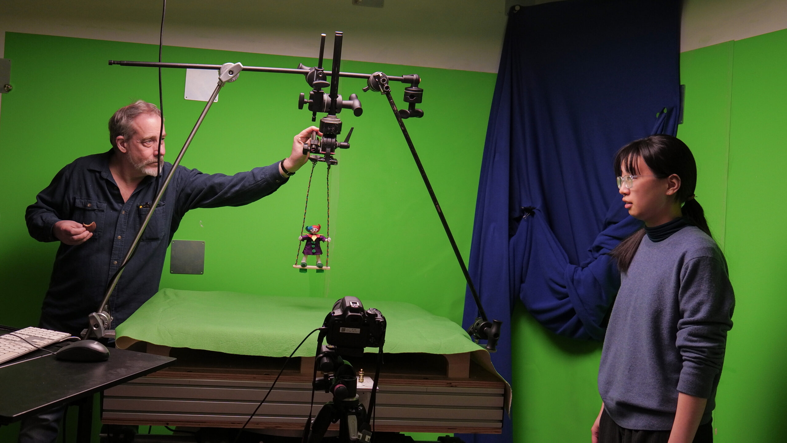A man with a beard is operating a camera setup in a studio with a green backdrop. He is holding a marionette puppet suspended from a hovering rig. Next to him, a young person with glasses and dark hair is observing the setup while standing on the right side. A computer keyboard is placed on a table in the foreground.