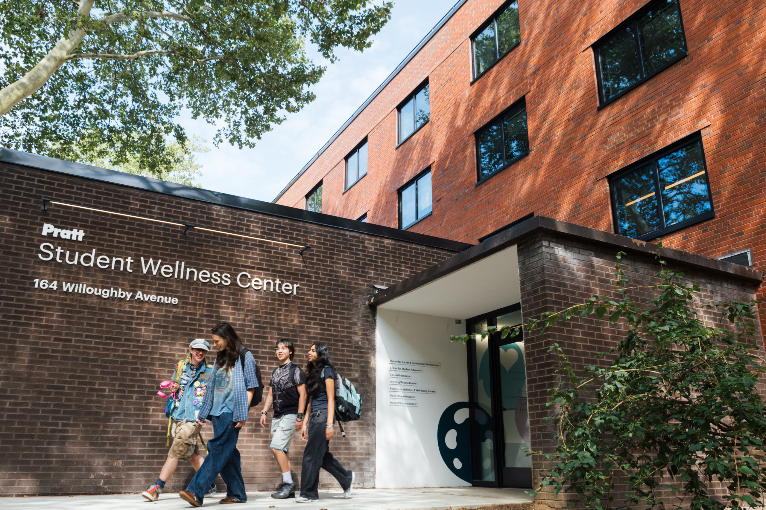 A group of four students walk towards the entrance of the Pratt Student Wellness Center, which features a modern brick facade. The center is located at 164 Willoughby Avenue, with trees providing shade in the background. One student wears a colorful outfit with patches, while others are dressed casually, and one carries a backpack. A sign with the center's name is visible next to the entrance.