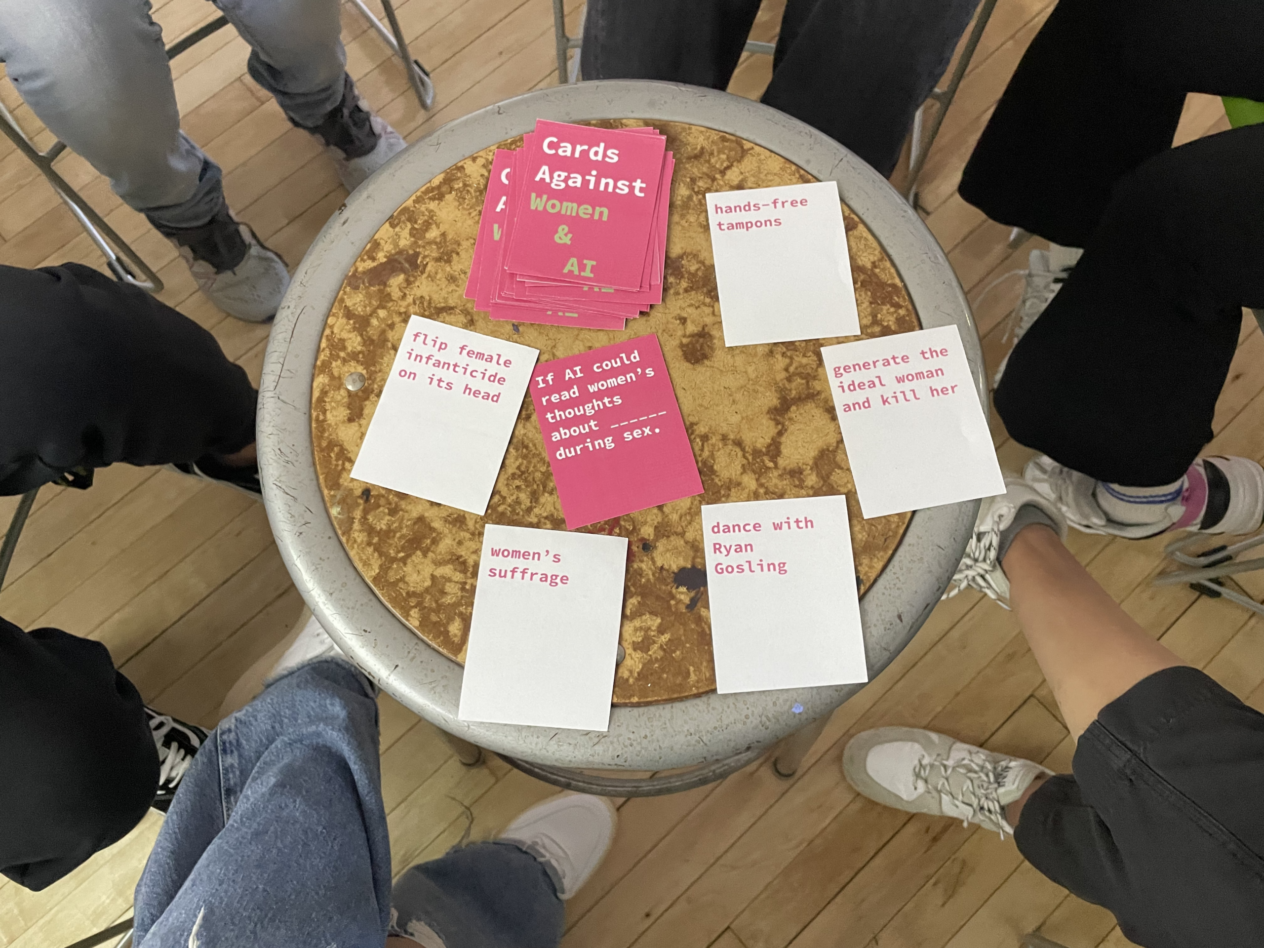 A circular table with a textured surface holds a stack of pink cards titled "Cards Against Women & AI." Surrounding it are several white cards with phrases such as "flip female infanticide on its head," "women's suffrage," and "dance with Ryan Gosling." The lower parts of people's legs and shoes are visible around the table, suggesting a group activity. The setting has a wooden floor.