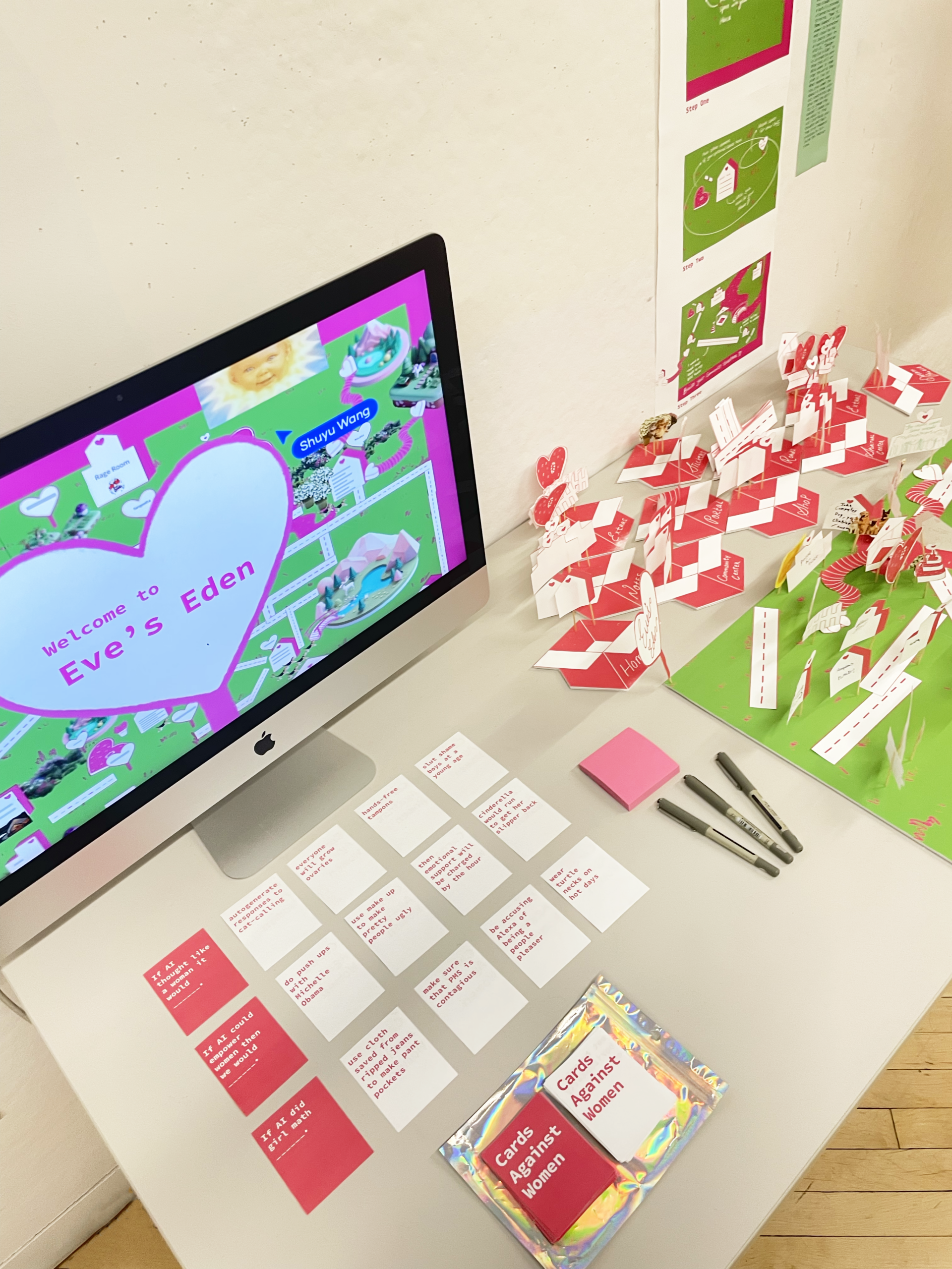 A computer monitor displaying a vibrant pink and green digital landscape titled "Welcome to Eve's Eden." Nearby, various printed cards and notes in red and white with text are arranged on a white table, along with a stack of colorful cards labeled "Cards Against Women." A pink sticky note and three markers are also on the table, with a decorative item in the background featuring small signs and illustrations.