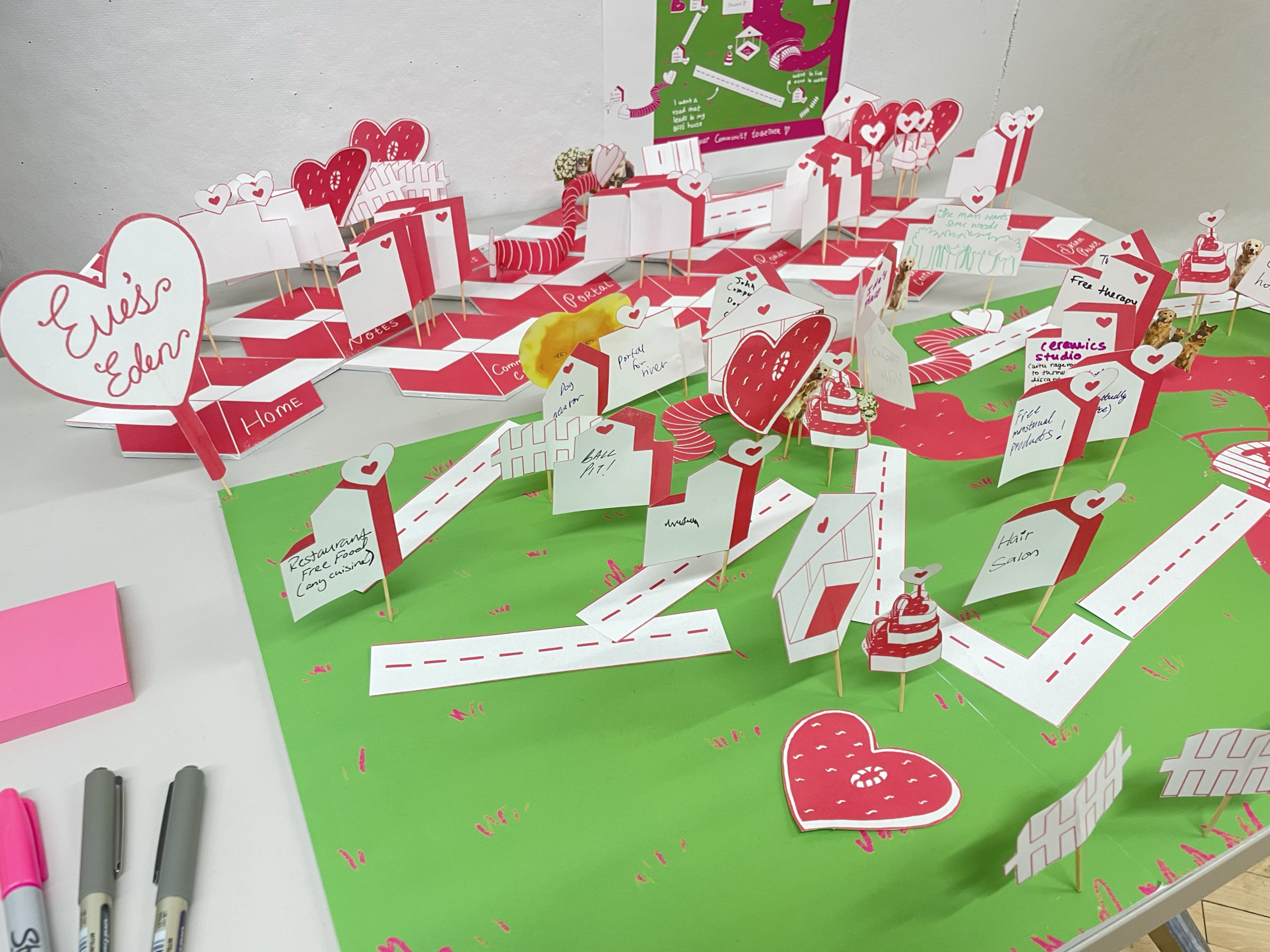 A colorful three-dimensional model featuring a green base and various red and pink pop-up structures resembling buildings, hearts, and signage. The scene includes labels for different locations such as a restaurant, hair salon, and ceramics studio, along with roads illustrated in white. Small decorations resembling cakes and other playful elements are scattered throughout, contributing to a whimsical atmosphere.