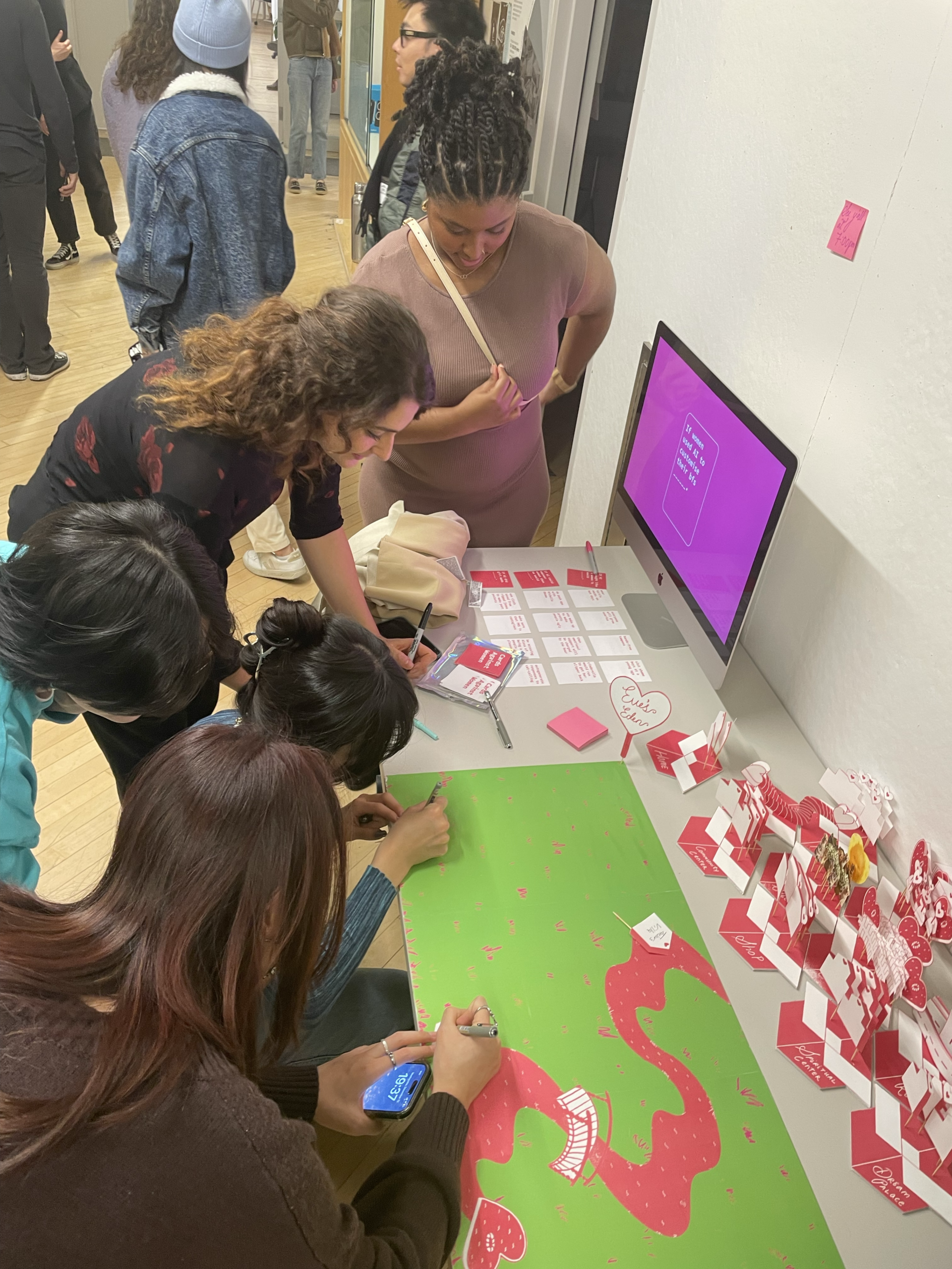 A group of four people is engaged in an activity at a table with a green poster and various printed materials. Two individuals are drawing on the poster, while another observes. A computer displays a vibrant pink screen near the table, which is also adorned with small cards. The background shows other attendees in a gallery-like setting.
