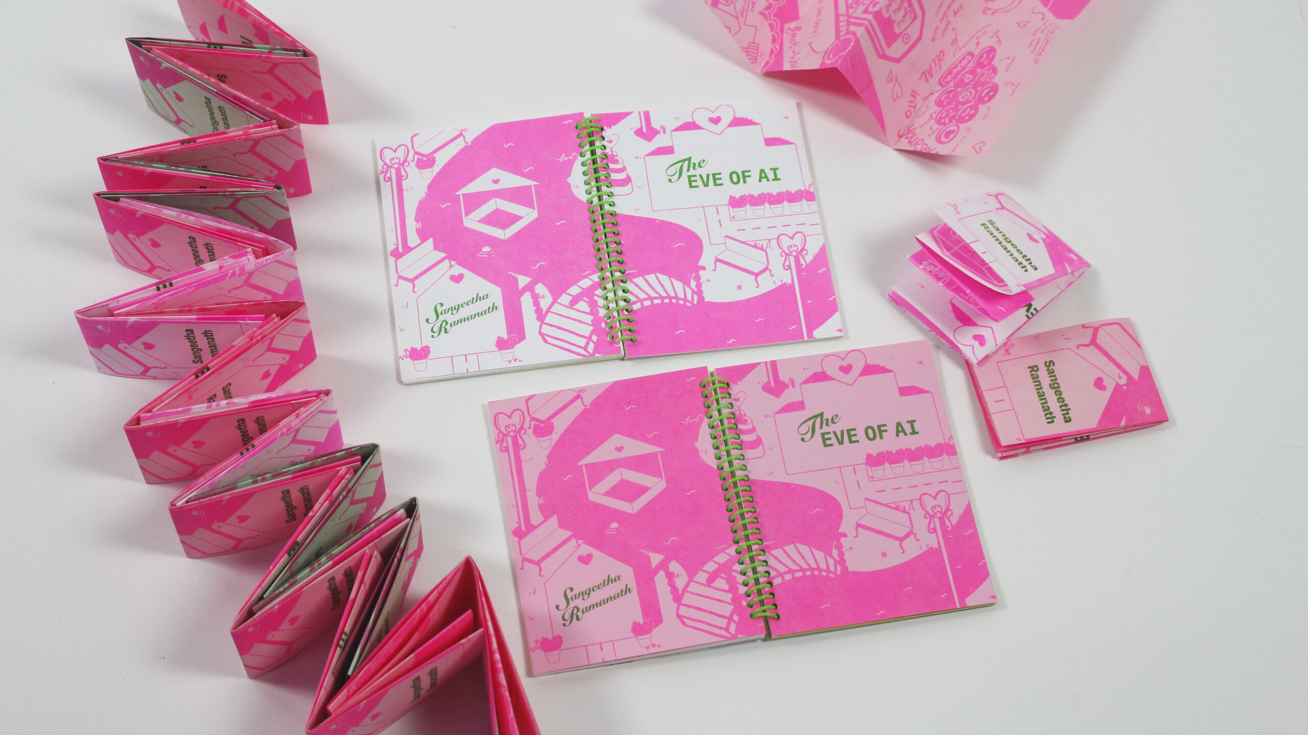 A collection of colorful pink booklets and a larger spiral-bound book titled "The Eve of AI" by Sangeetha Ramanath. The books feature playful, illustrated designs with hearts and other small graphics. The smaller booklets are arranged in a zigzag pattern, while the larger book lies open to reveal its illustrated pages. The overall presentation conveys a vibrant and artistic theme.