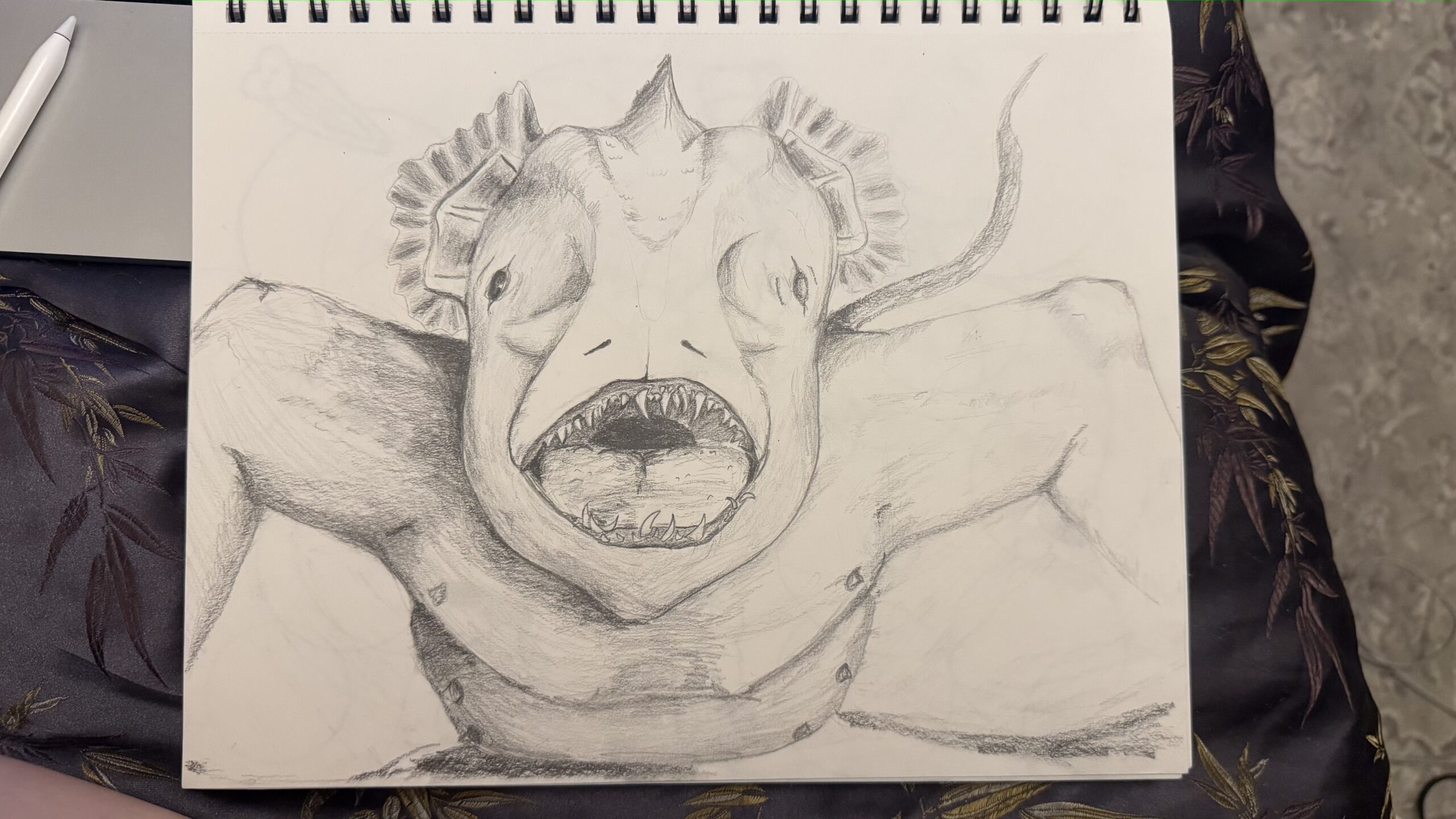 A detailed pencil sketch of a monstrous creature resembling a blend of a fish and a human. The creature has a wide, toothy mouth, large eyes, and fins on its head, with muscular arms and a body positioned in a confrontational stance. The background features a textured surface, and the sketch is situated on a patterned fabric.