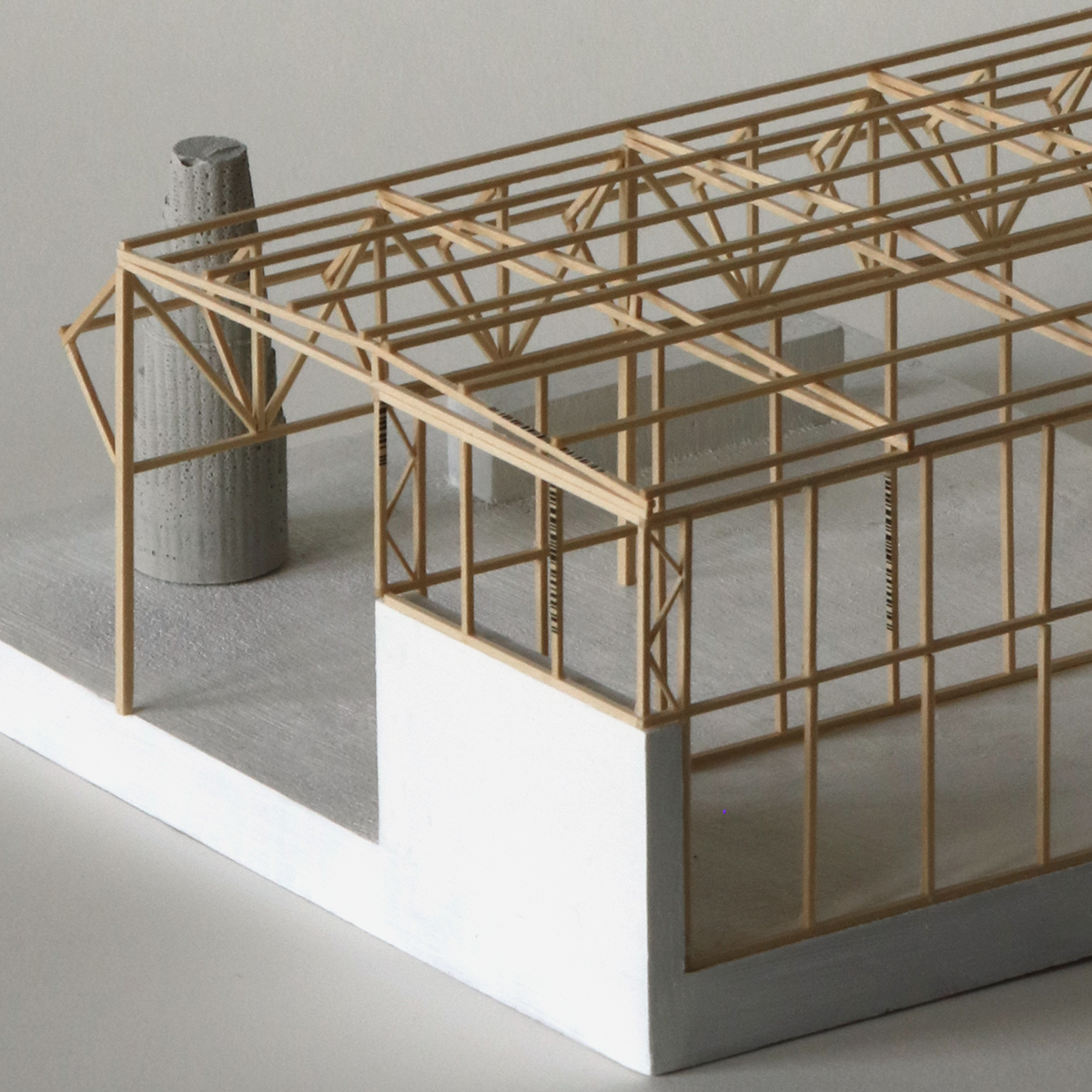 A model of a building structure featuring a wooden frame and concrete elements. The structure has a partially exposed interior with a gray base and white sections. A cylindrical concrete feature stands near one end of the model. The overall design reflects a minimalist architectural style.