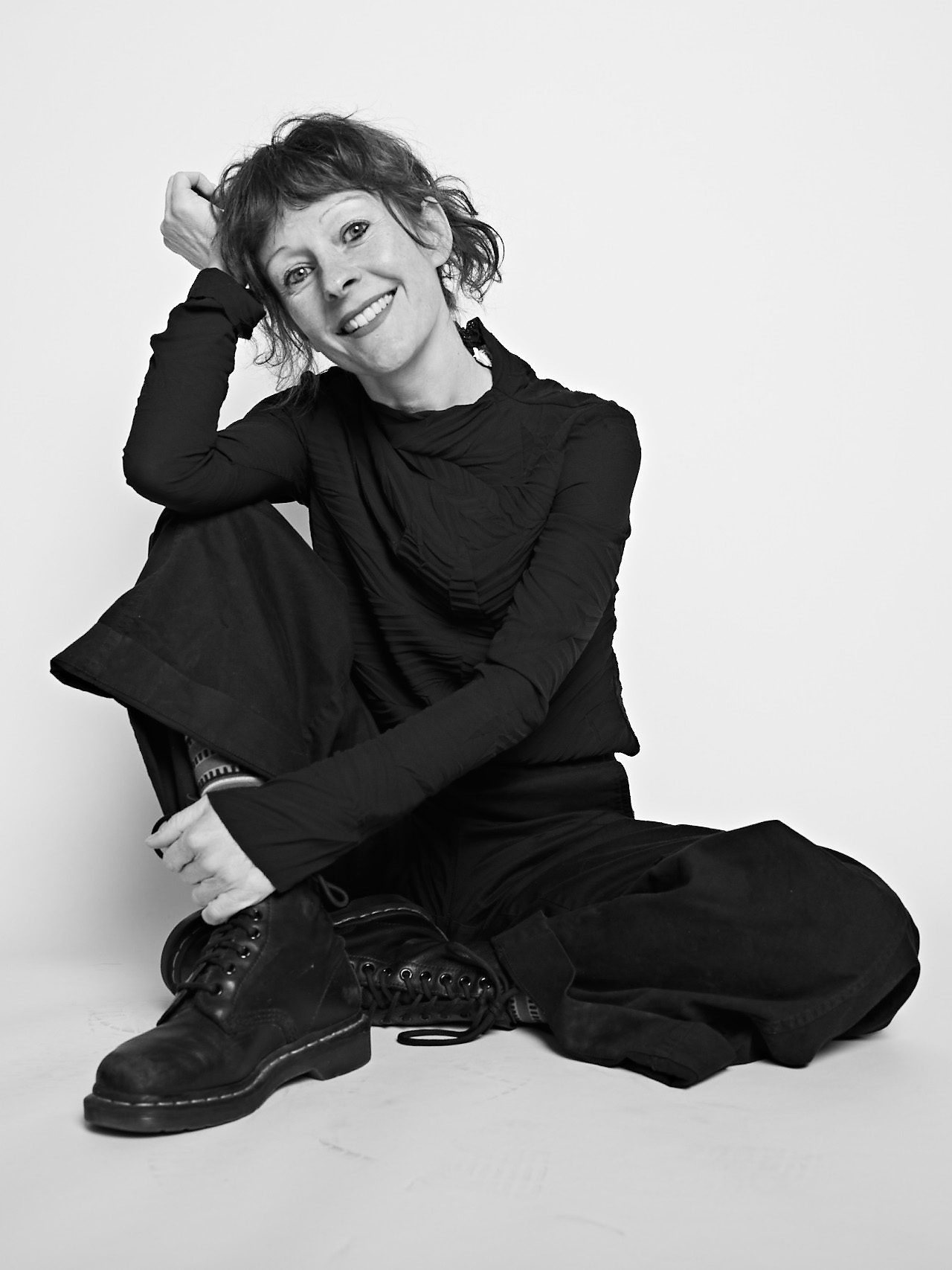 Black and white photo of smiling woman seated on the floor. She has her legs crossed and is dressed in black pants and shirt and is wearing black work boots.