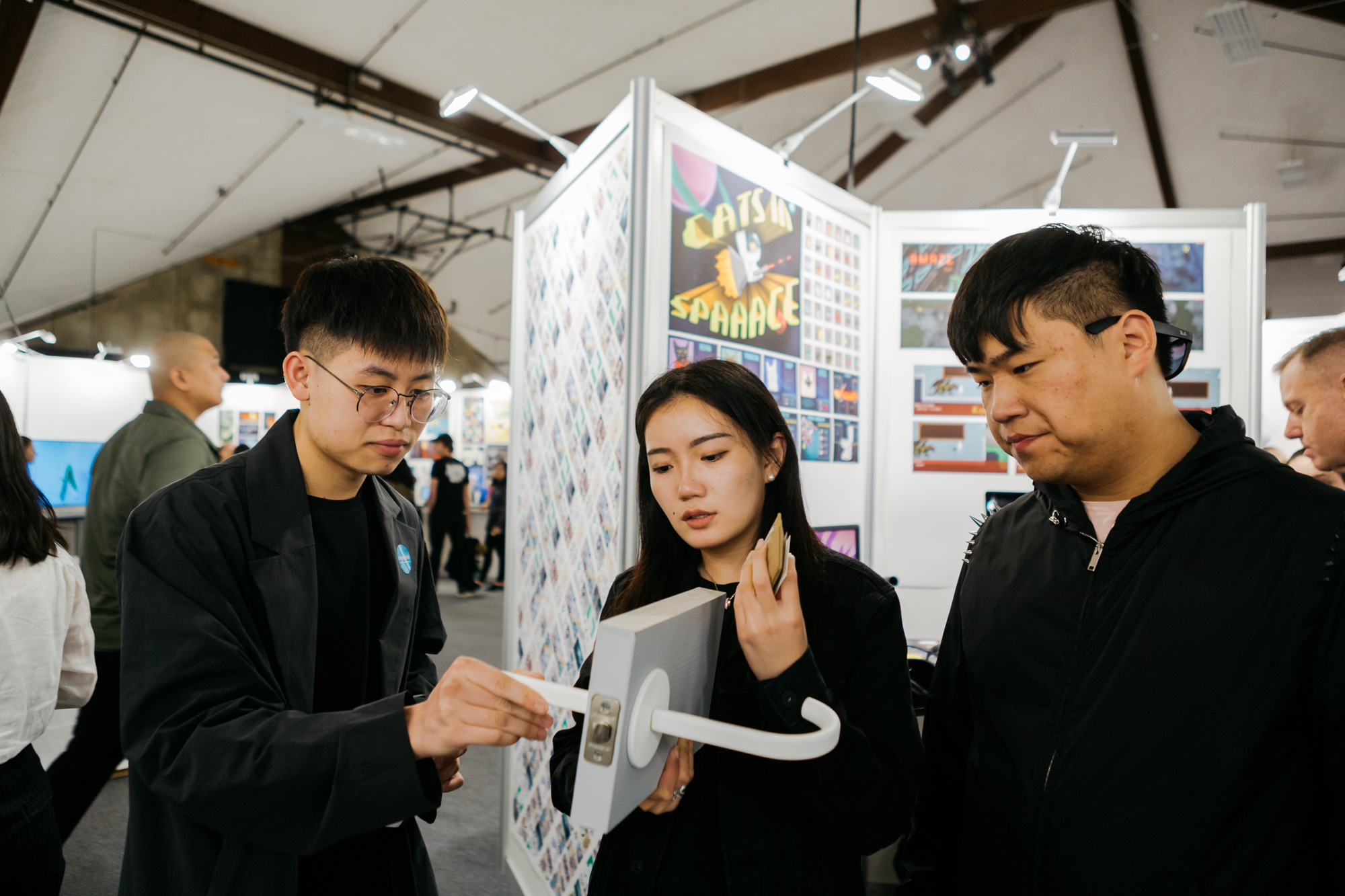 A group of three young adults is gathered at an art exhibition. One person, wearing glasses and a black jacket, is demonstrating a device to a woman holding a card. She appears focused on the device, while another man in a black jacket watches attentively. Behind them is a display featuring colorful artwork, including a sign that reads "Cats in Space." The exhibition environment has a modern aesthetic with ample lighting.