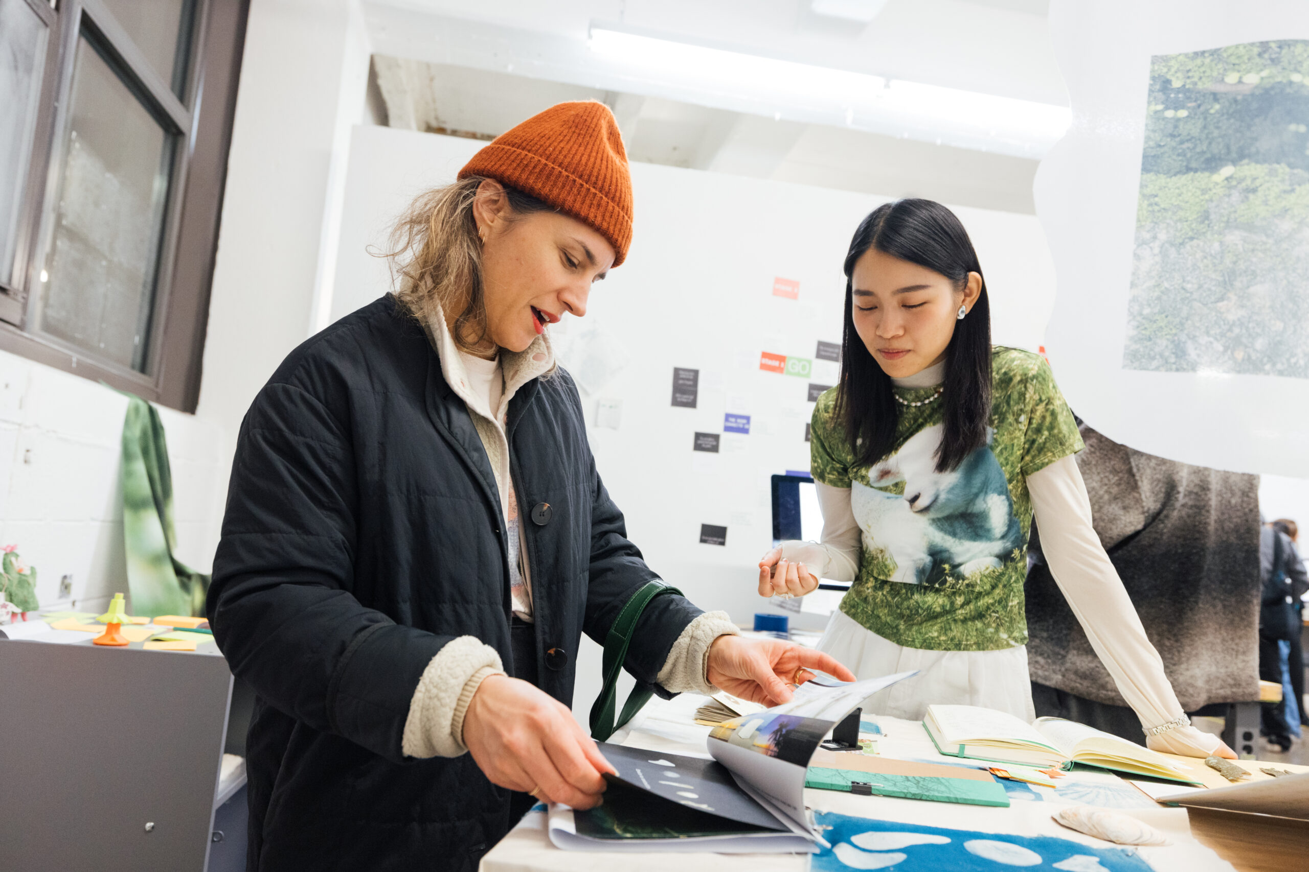 Two individuals are engaged in discussion while looking at a book on a table filled with various materials. One woman, wearing an orange beanie and a dark jacket, appears surprised or intrigued. The other woman, dressed in a green graphic t-shirt overlaying a white long-sleeve top, stands beside her, observing the book. The background shows a workspace with colorful notes and materials scattered around.