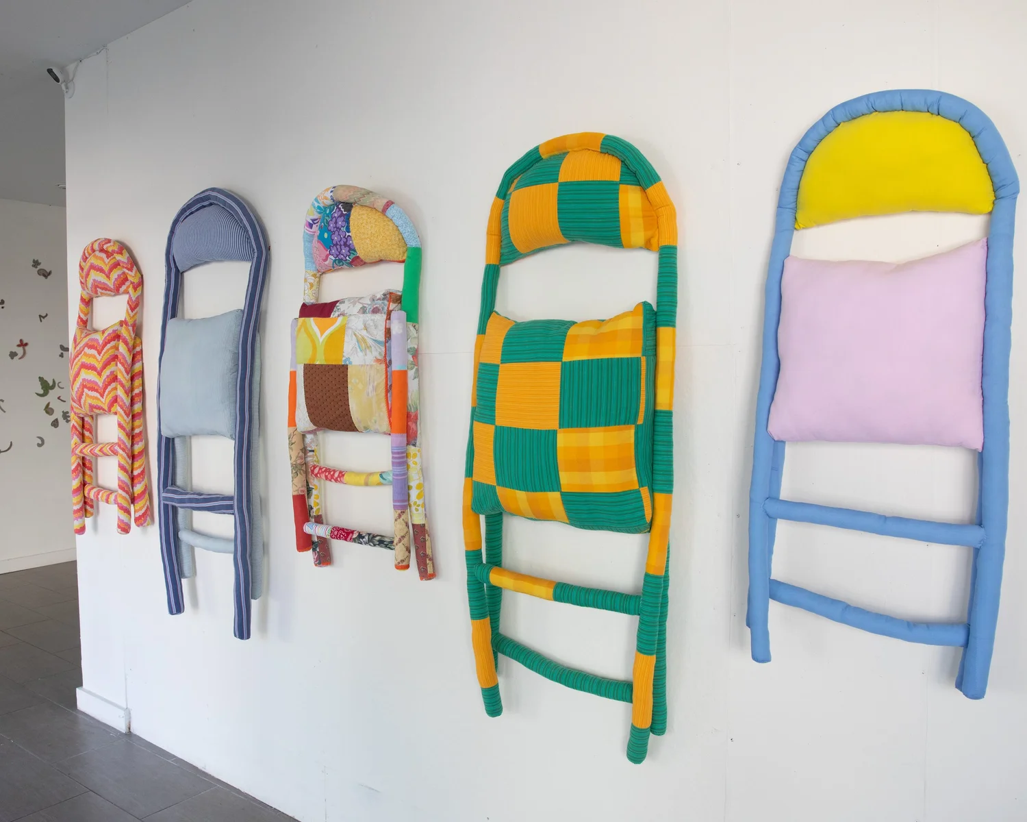 Five decorative art pieces resembling ladders are mounted on a white wall. Each ladder has a unique pattern and color scheme, featuring wrapped fabrics in bright hues like red, purple, green, yellow, and orange, with padded sections resembling cushions. The artistic display blends various textiles, creating a playful and vibrant aesthetic.