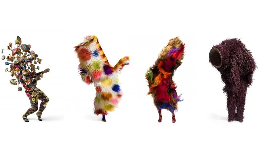 Four figures are dressed in vibrant, extravagant costumes. The first figure has a body covered in a floral pattern, with various musical instruments emerging from their head and shoulders. The second figure is enveloped in a colorful, fluffy outfit resembling a rainbow. The third figure displays a similar fluffy costume with bright colors. The fourth figure appears to be wearing a dark, furry costume, resembling a large silhouette with an empty head section.