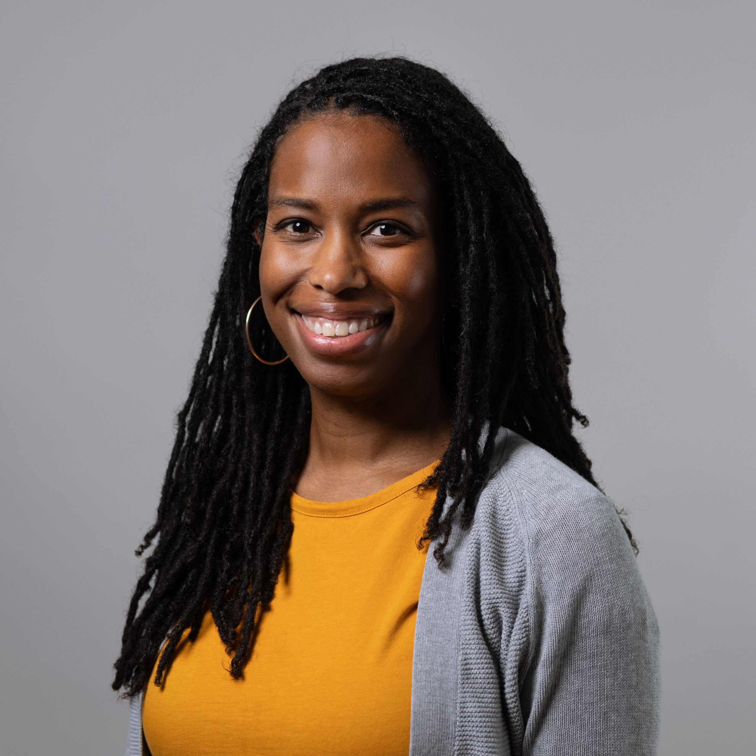 A smiling woman with long, dark locs wearing a mustard yellow shirt and a light gray cardigan. She has large hoop earrings and stands in front of a plain gray background.