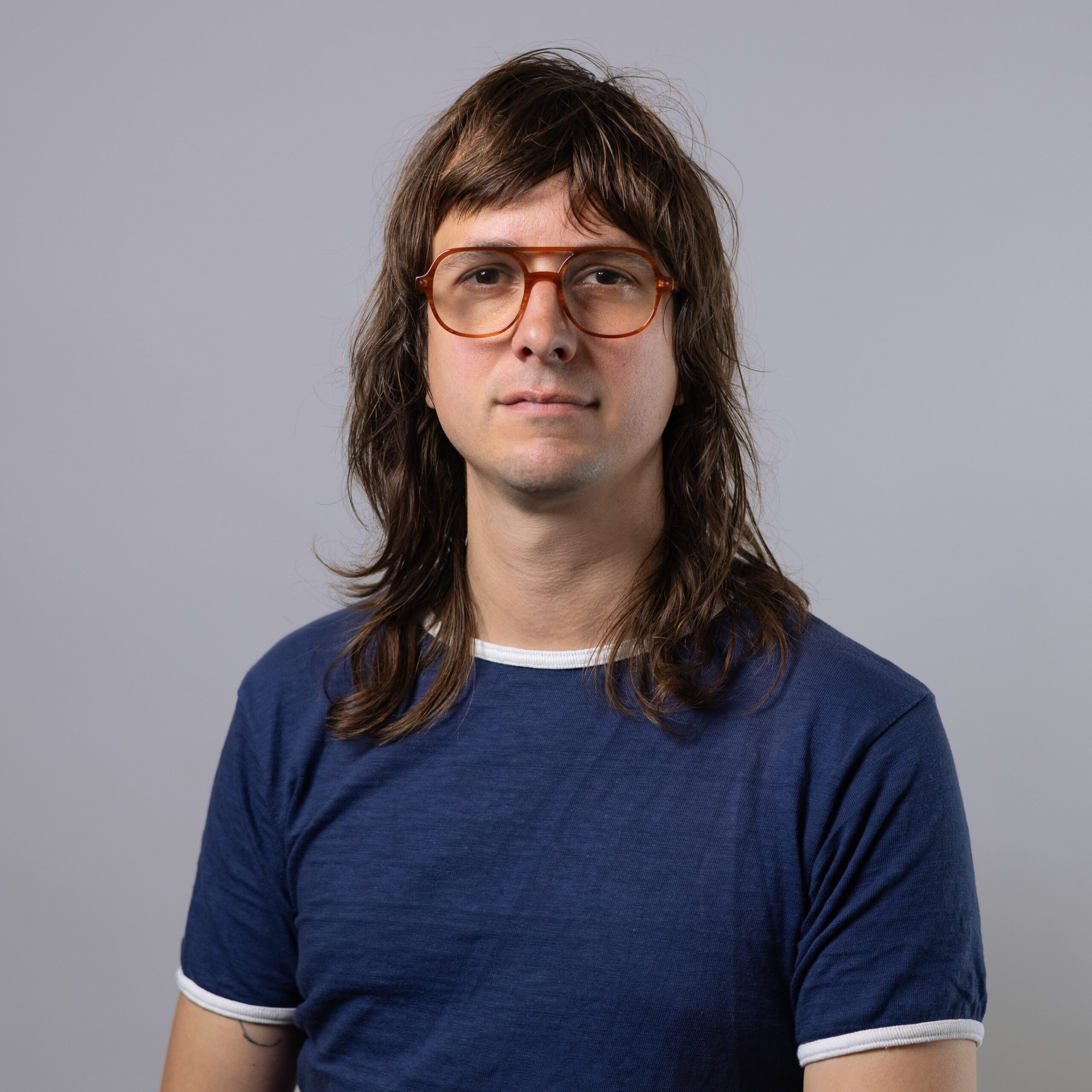 A person with long, wavy brown hair wears orange glasses and a blue t-shirt with white trim. They are facing forward with a neutral expression against a light gray background.