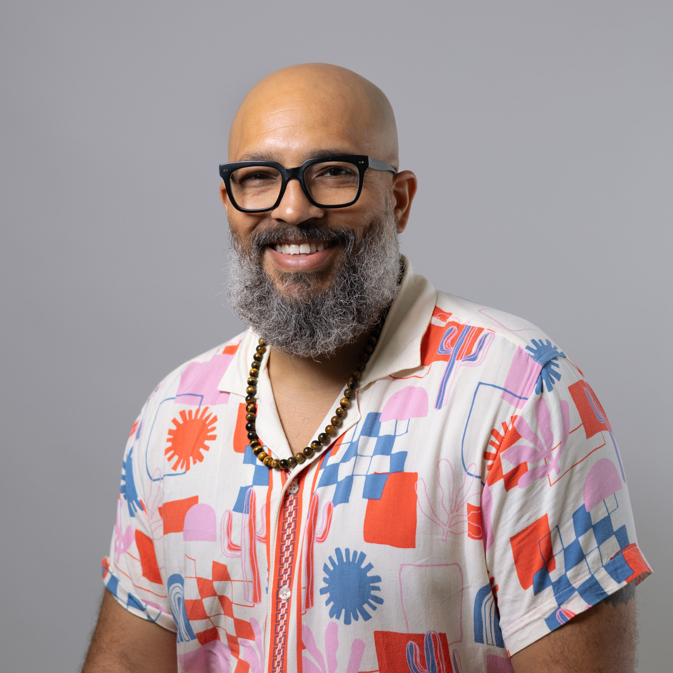 A smiling individual with a bald head and a full, gray beard is wearing black glasses and a colorful, patterned short-sleeve shirt with various shapes in red, blue, pink, and white. They are also wearing a beaded necklace and are posed against a plain light gray background.